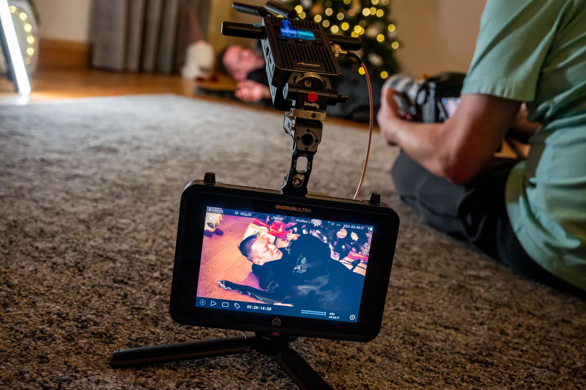 A camera monitor on a tripod shows a touching scene of a man hugging a dog. To the right, a person is filming on a carpeted floor, creating a warm, cozy atmosphere.