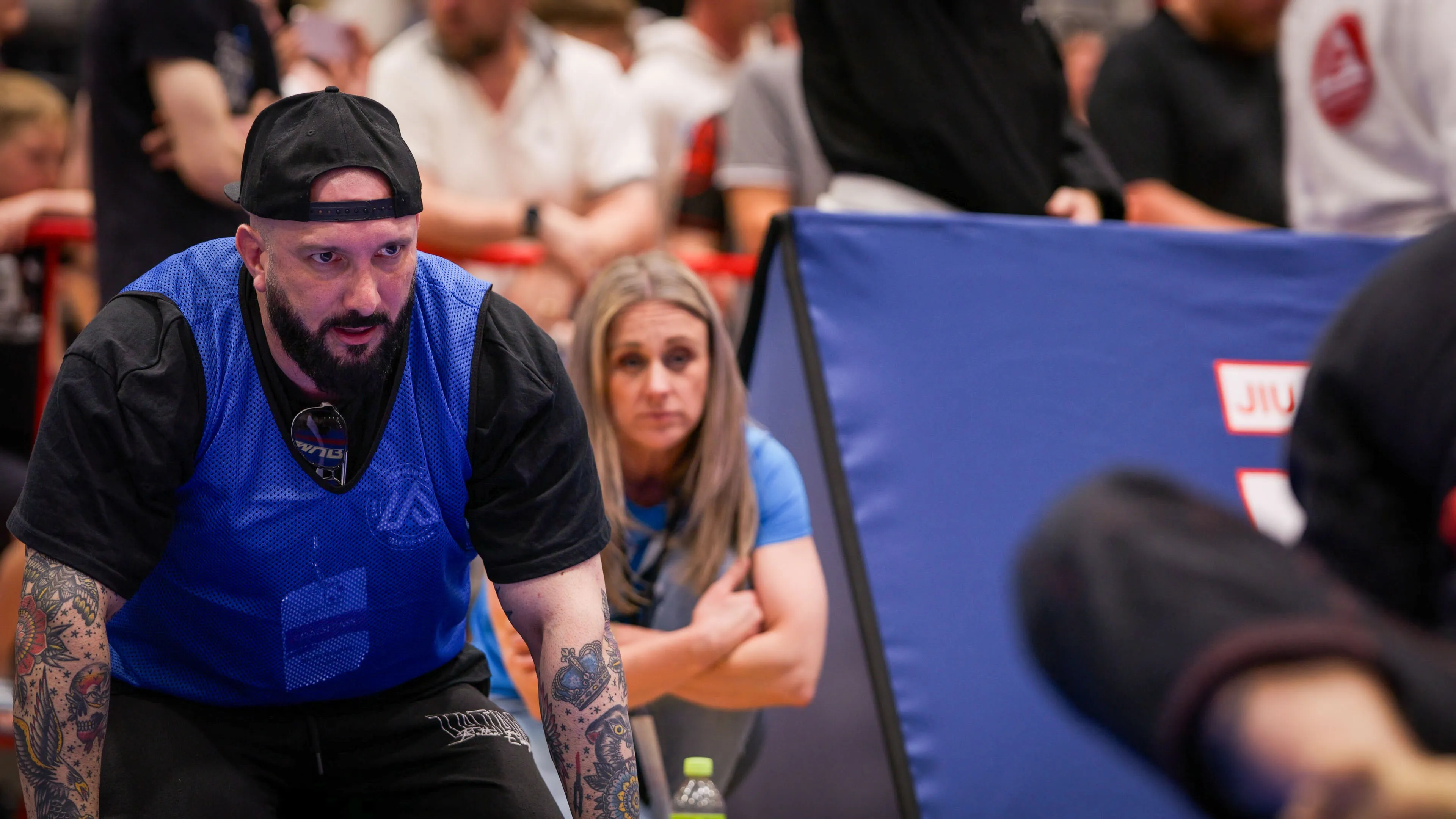 A focused, tattooed man in a blue sports vest crouches at a Jiu-Jitsu event, with a woman attentively watching in the background amid a lively crowd.