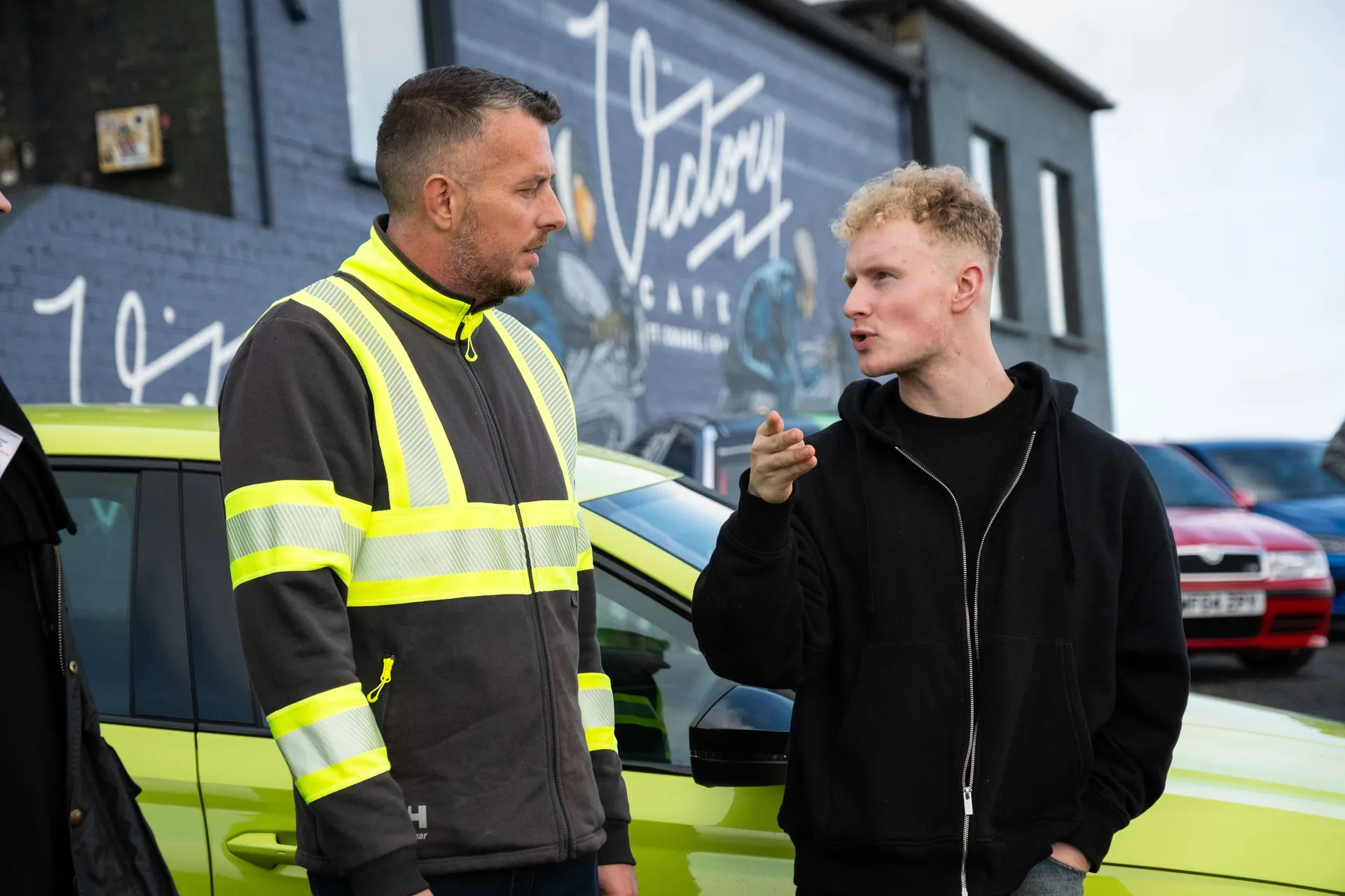 A man in a high-visibility jacket and another in a black hoodie converse next to a bright green Skoda car. A mural reading "Victory" is on the building behind them.