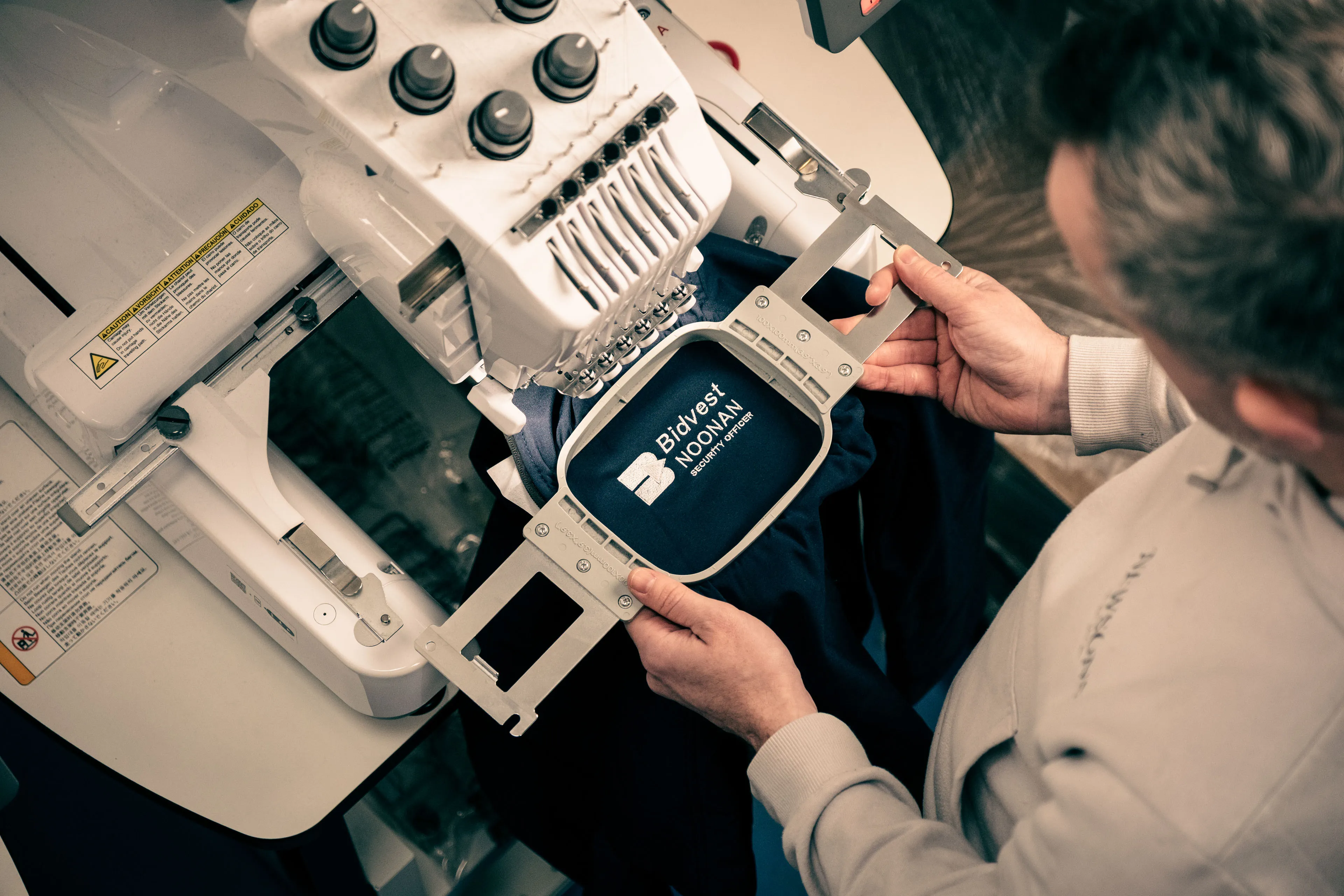 A person adjusts a hoop on an embroidery machine featuring a navy garment labeled "Bidvest Noonan Security Officers."