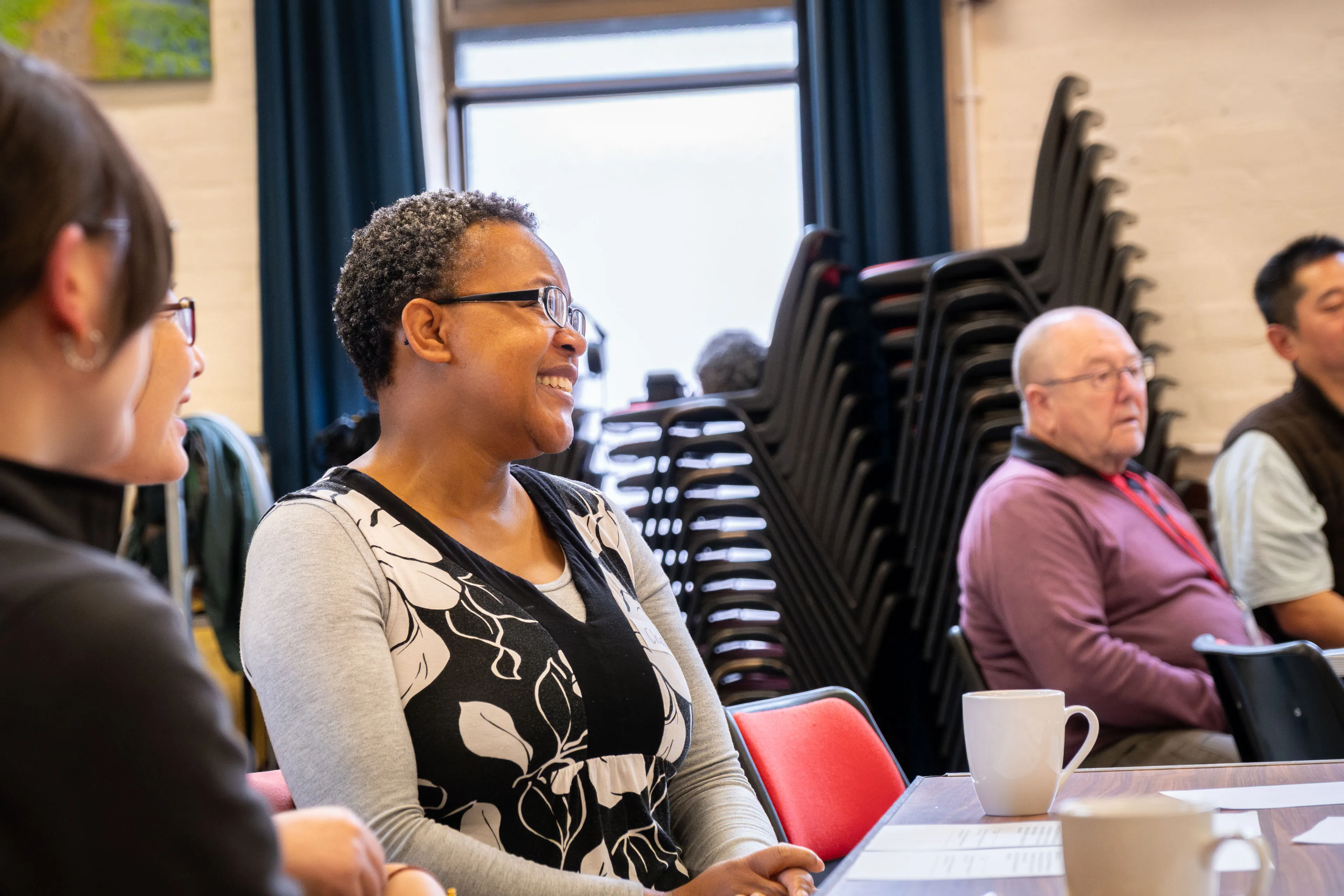 A group of people sit at a table in a bright room, smiling and engaged. Stacked chairs are visible in the background, creating a casual, warm atmosphere.