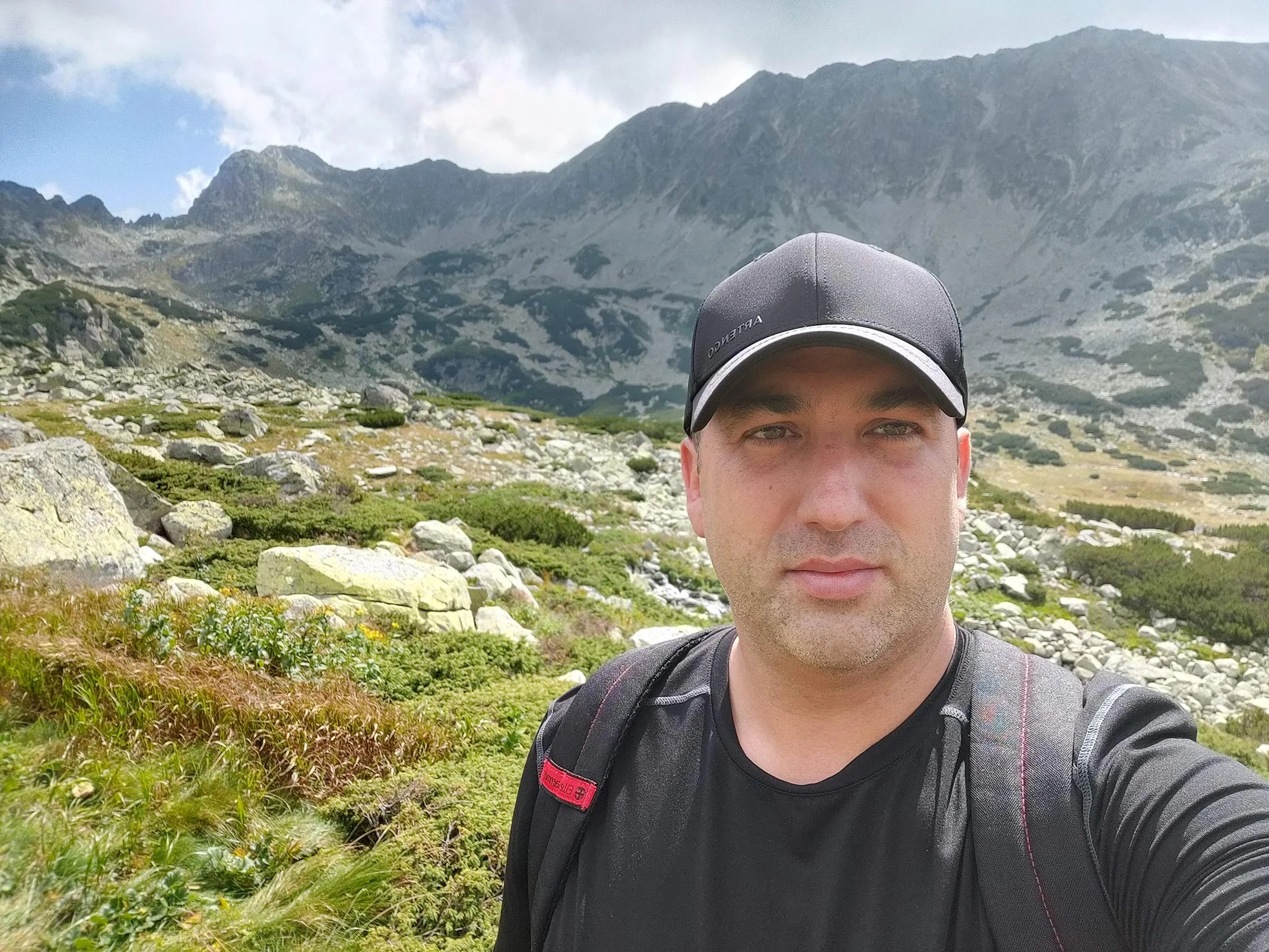 Claudiu Danila taking a selfie on a mountain trail wearing a black cap and backpack, with a rocky mountainside and Eastern European landscape behind him