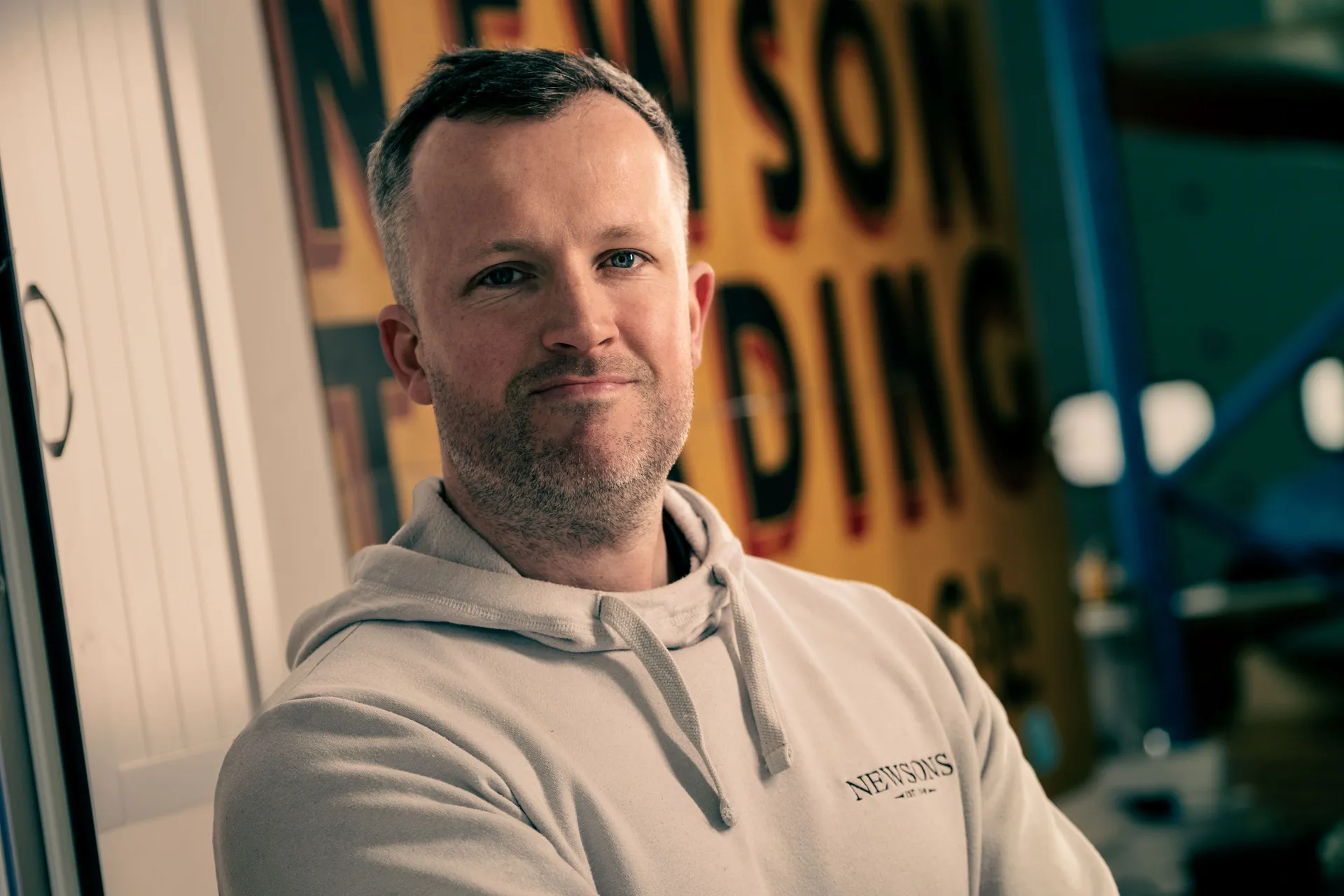 A man in a hoodie stands confidently with arms crossed. Behind him is a large, partially visible, yellow sign. The mood is relaxed and self-assured.