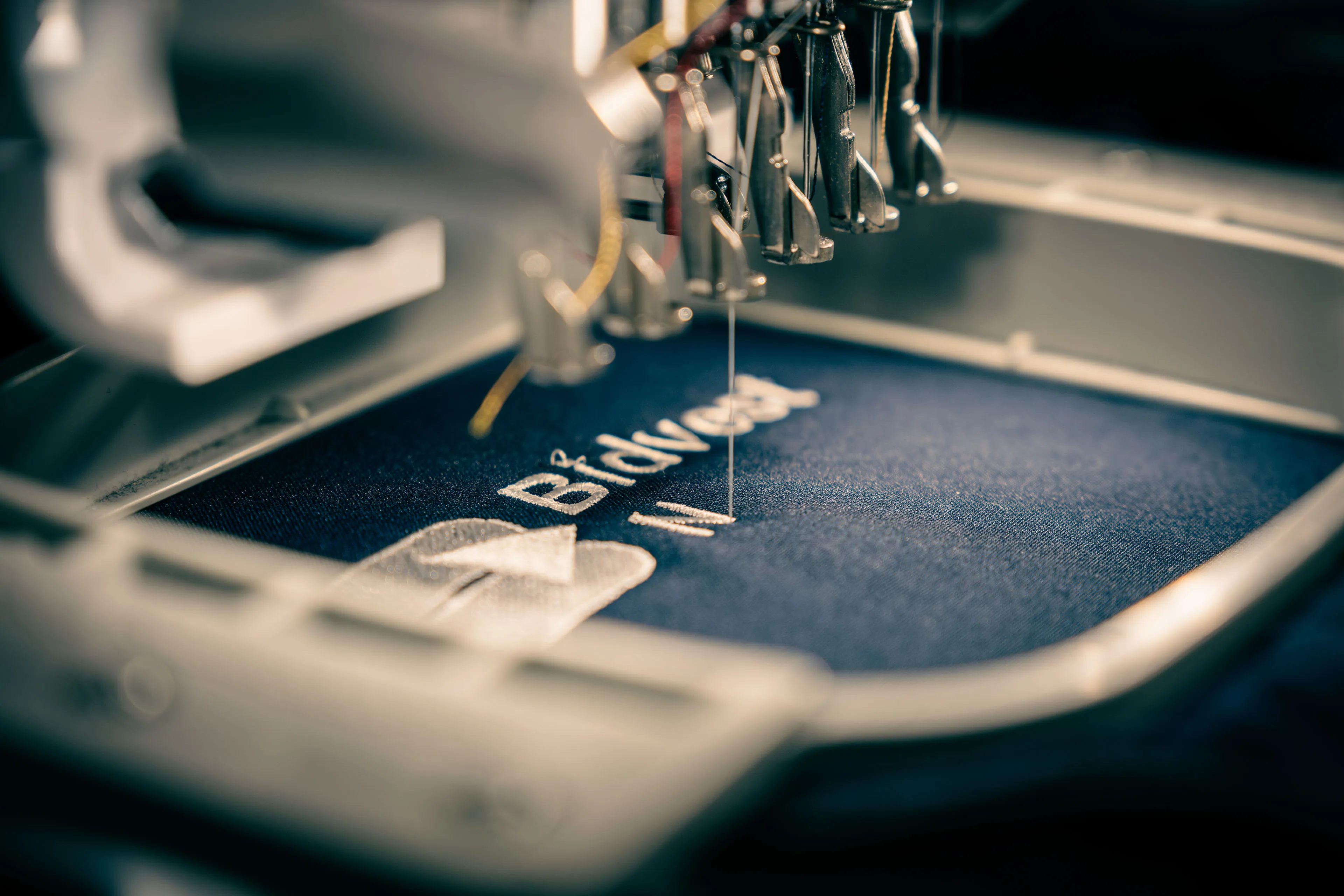 Close-up of a sewing machine embroidering white text and a logo on dark fabric. The image conveys precision and craftsmanship.