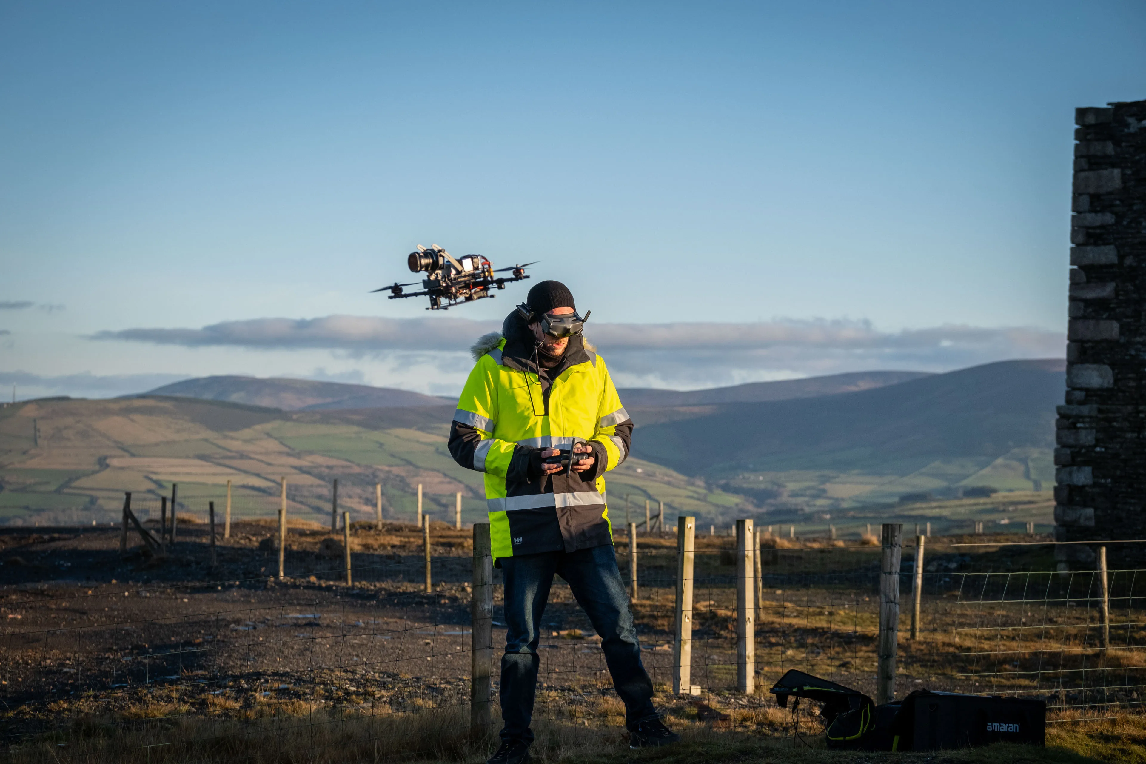 A man in a bright green/yellow jacket operates a fpv drone with a remote, standing on a grassy hilltop with a mountain landscape. The scene is calm and scenic.