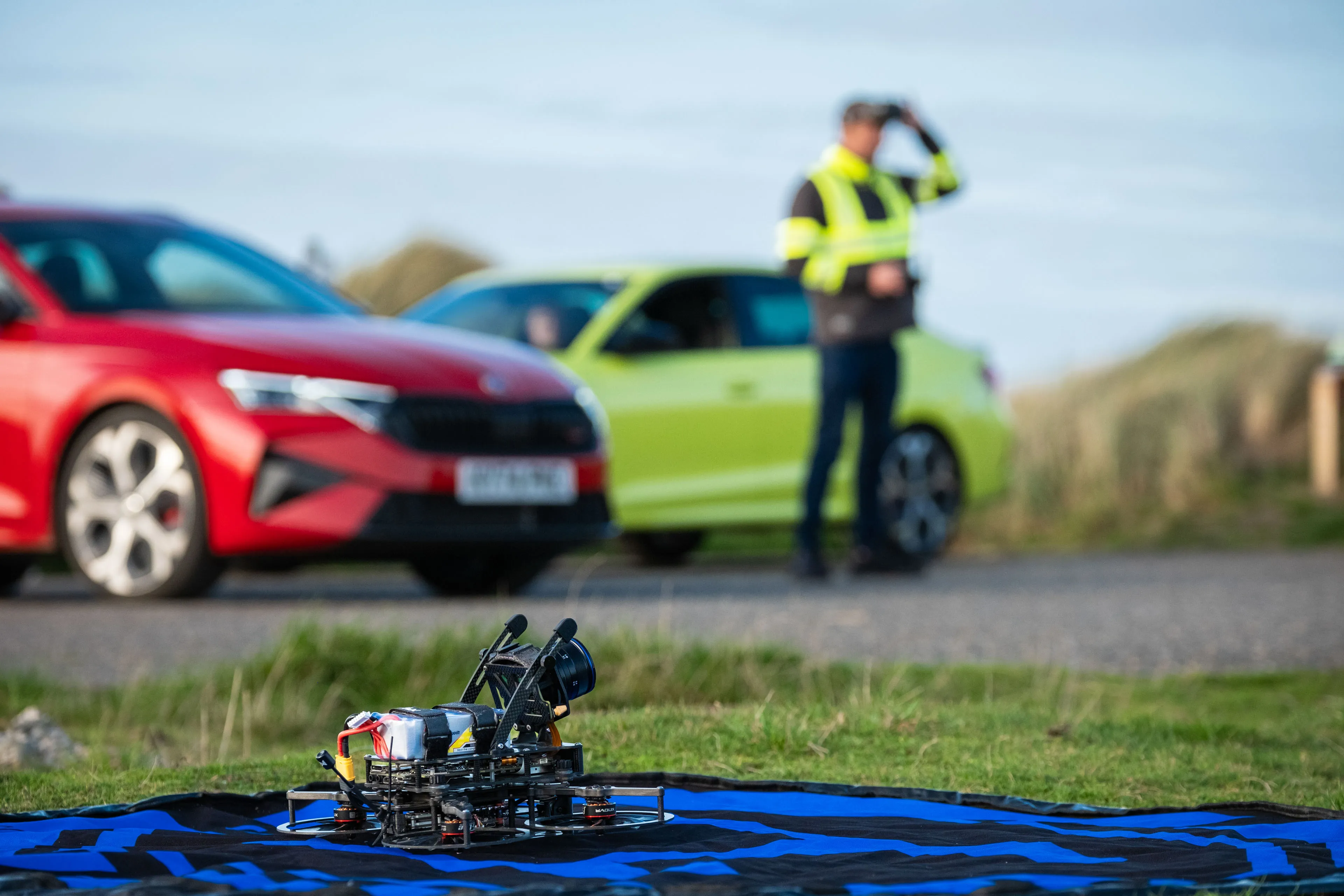A drone sits on a blue landing pad in the foreground, with a red and a lime green Skoda car parked nearby. A person in a high-vis jacket stands beside the cars.