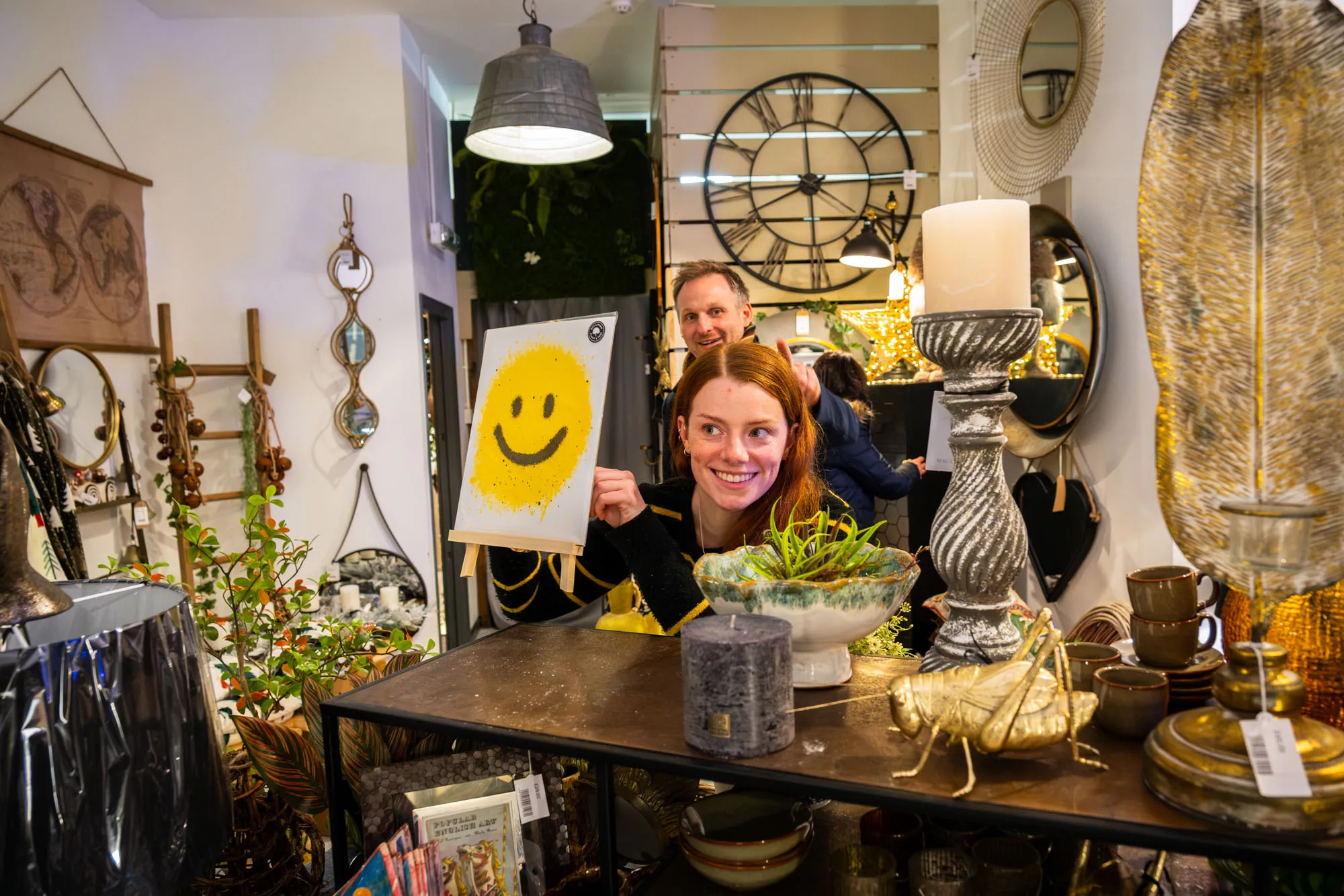 Woman holding a smiley face art print in a cozy, decorative shop filled with lamps, mirrors, and plants. She looks cheerful and inviting.