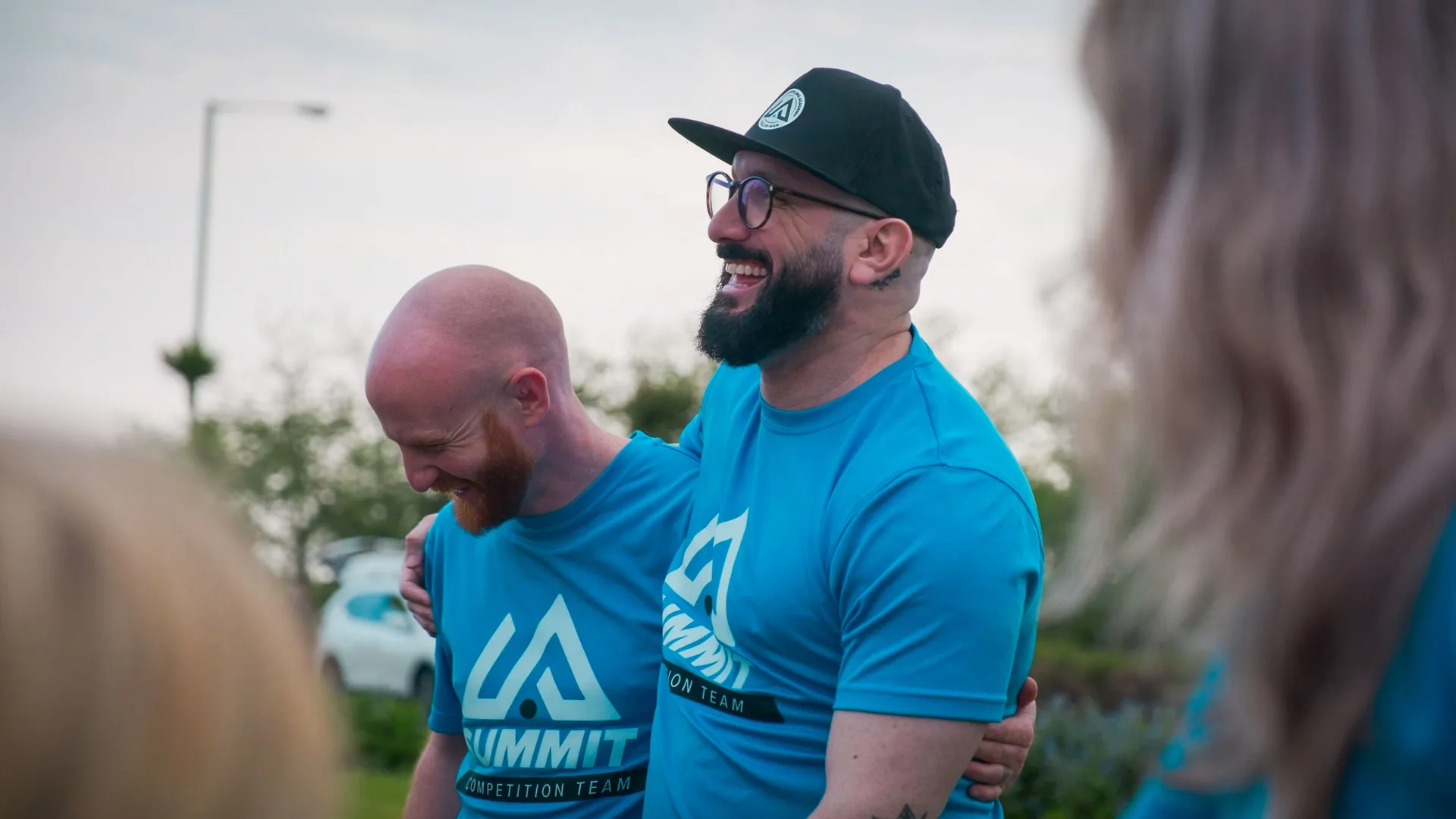 Two men in matching blue t-shirts with "Summit Competition Team" logos are laughing and embracing outdoors. The mood is joyful and relaxed.