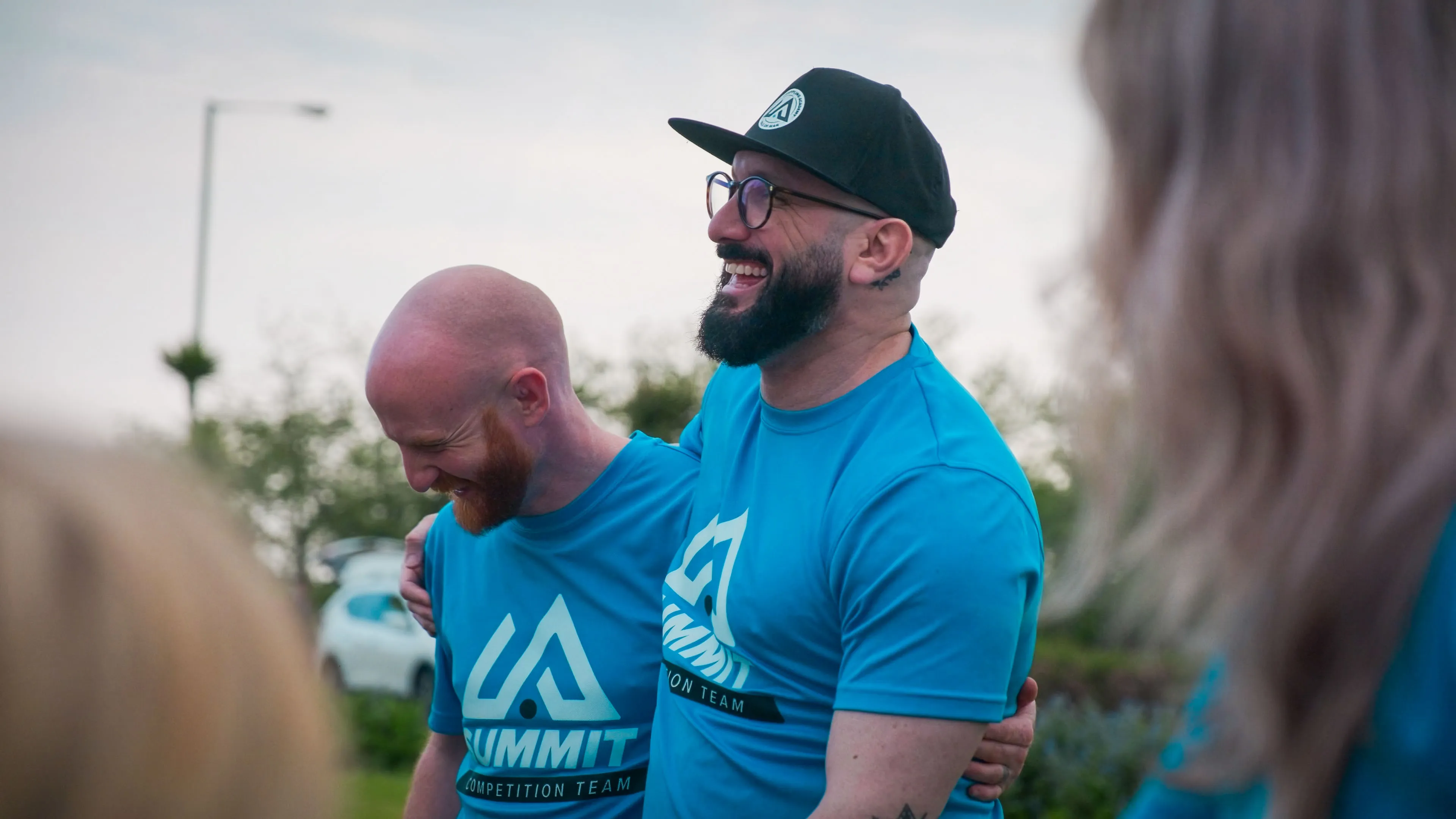 Two men in matching blue t-shirts with "Summit Competition Team" logos are laughing and embracing outdoors. The mood is joyful and relaxed.