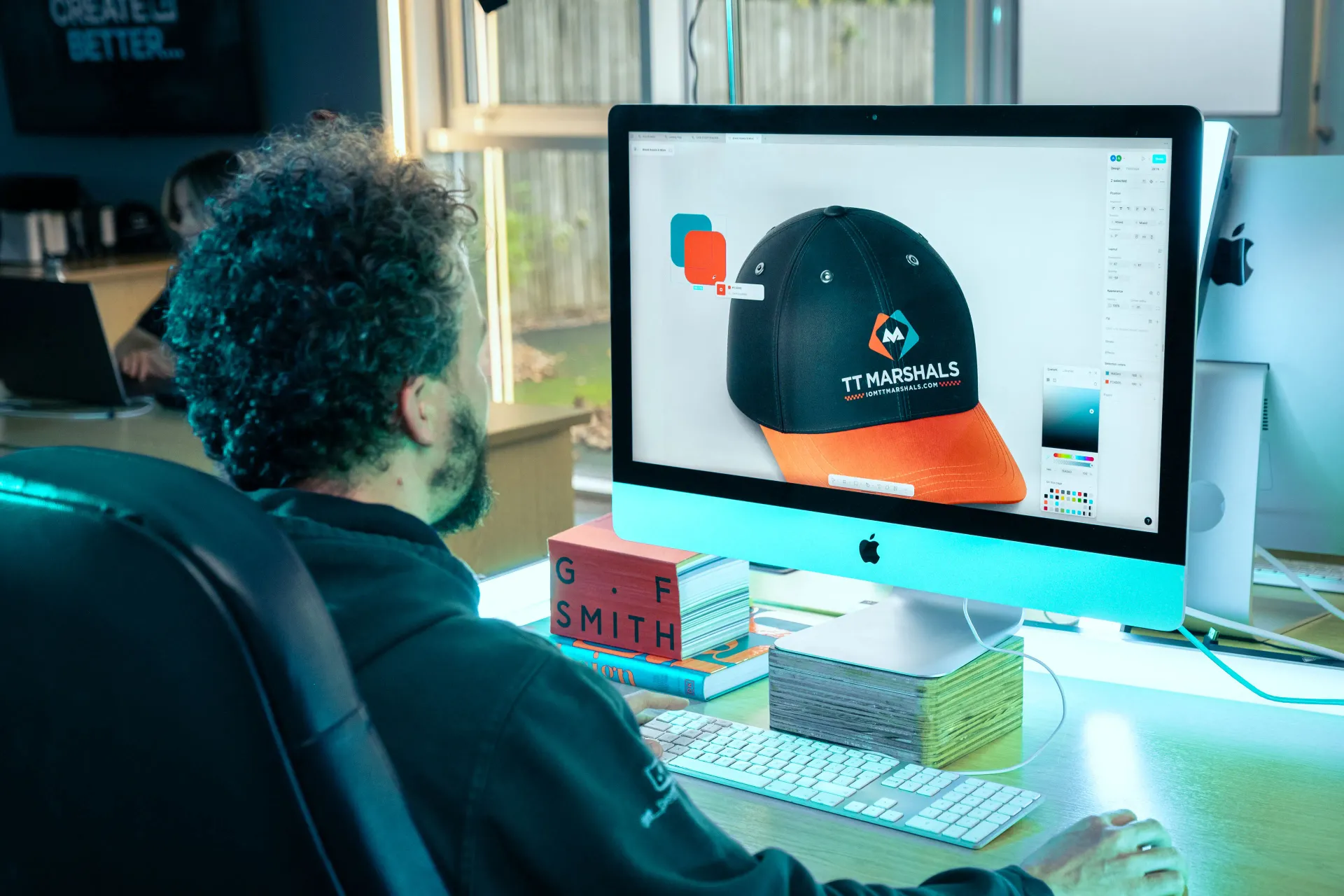 A DotPerformance designer working at an iMac in the Douglas office, designing a TT Marshals branded cap with the logo and iomttmarshals.com URL