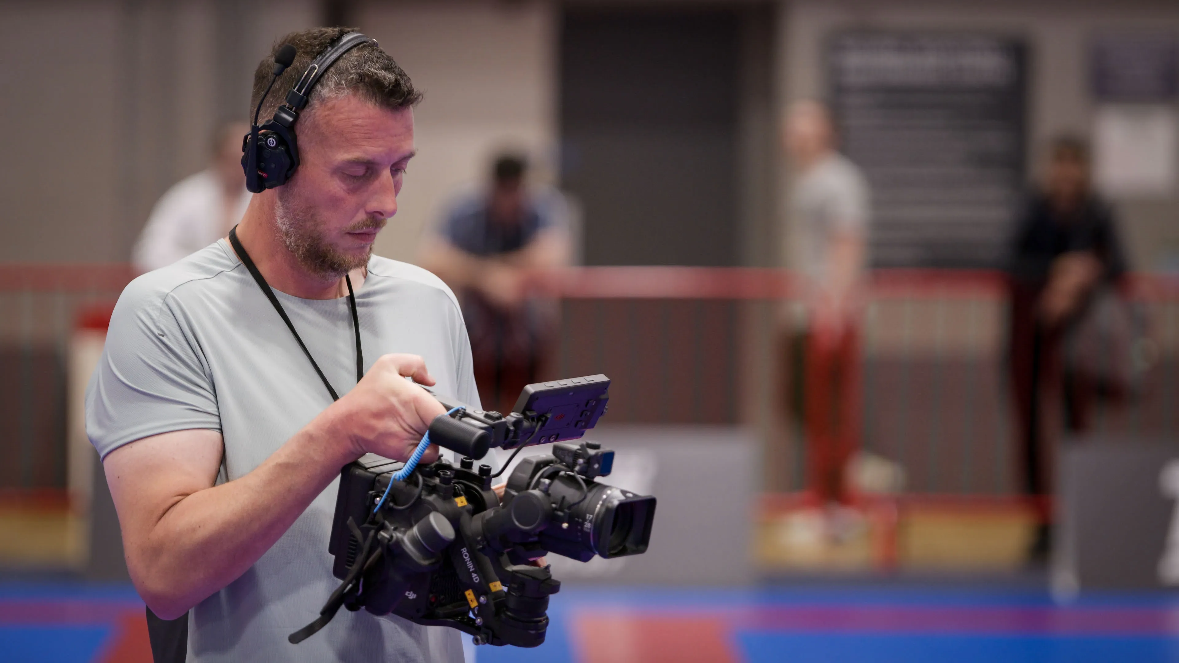 A man in a grey shirt and headphones carefully operates a professional video camera in a gym setting. The background shows blurred figures and gym equipment.