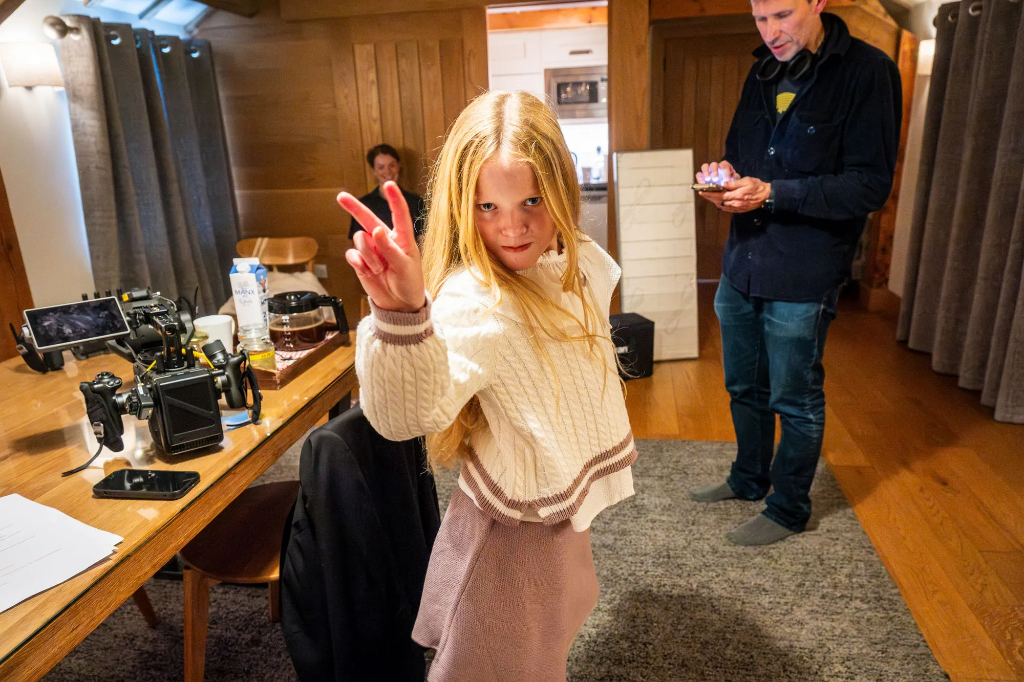 Young girl with long blonde hair flashes a peace sign, wearing a cozy sweater. A man stands nearby in a wooden room with cameras on a table, creating a casual and lively atmosphere.