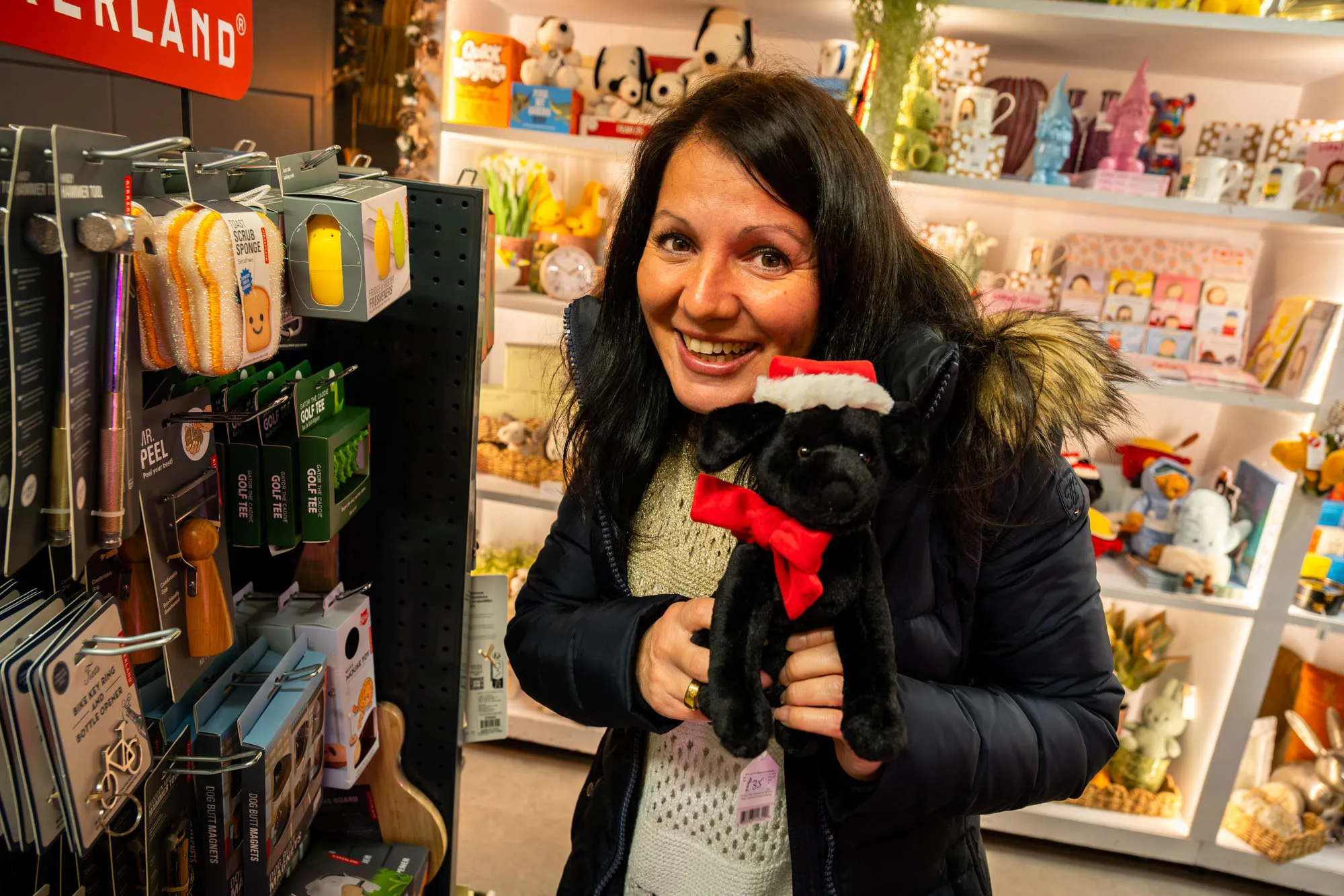Smiling woman in a shop holding a plush dog wearing a Santa hat. Shelves are filled with colourful toys and gifts, creating a festive atmosphere.