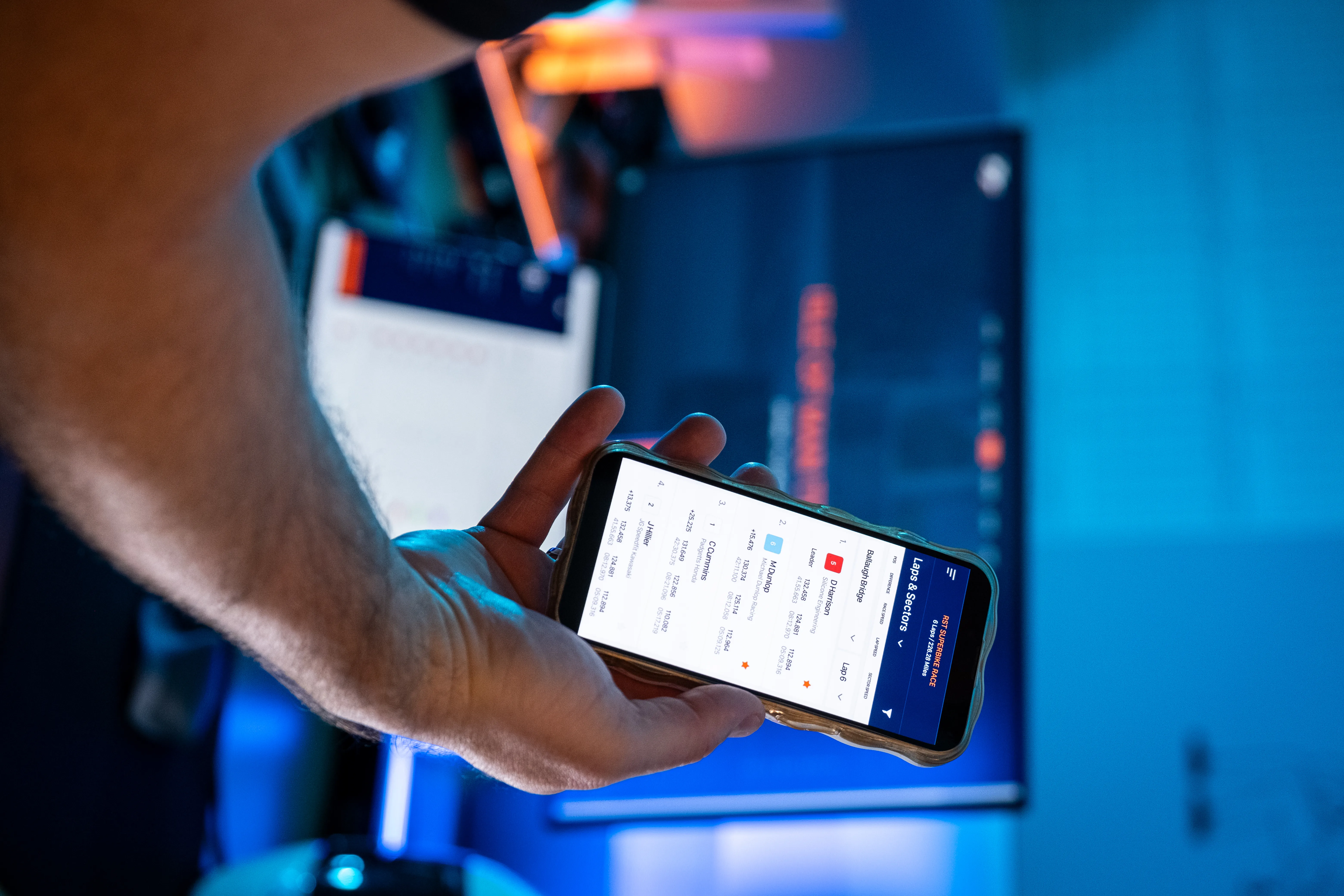 A person holds a smartphone displaying a finance app with stock listings. A computer monitor glows in the dimly lit room, creating a tech-focused atmosphere.