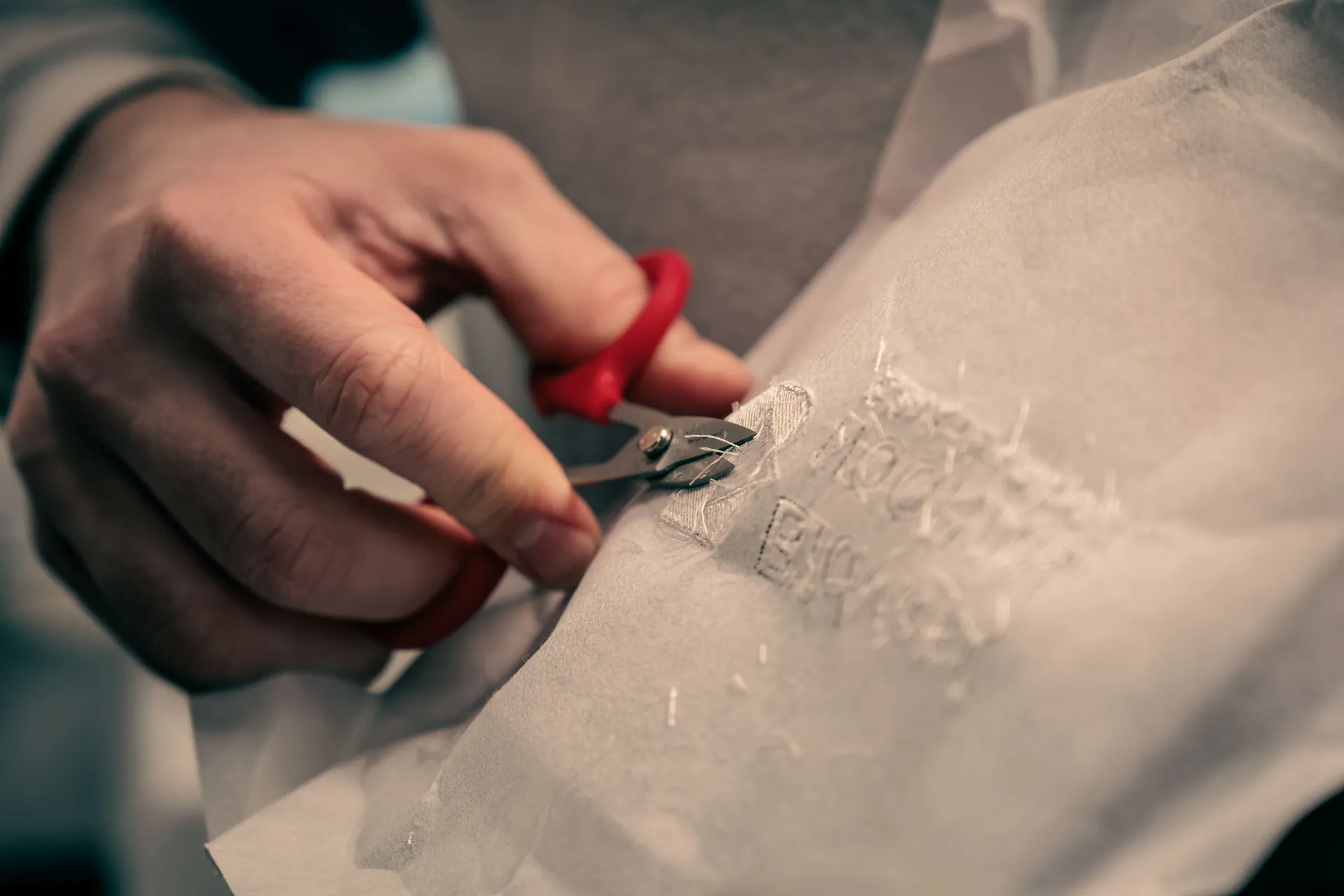 A hand carefully uses small red scissors to trim white material with stitched text. The focus is on precision and detail in fabric crafting.