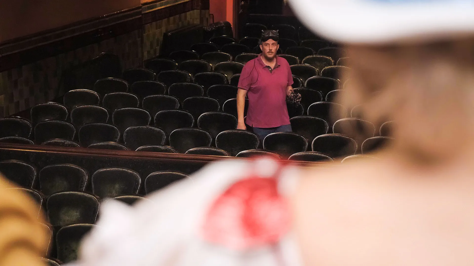 A man in a pink shirt stands among empty theater seats, looking forward. A blurred figure in the foreground watches him, creating a contemplative mood.