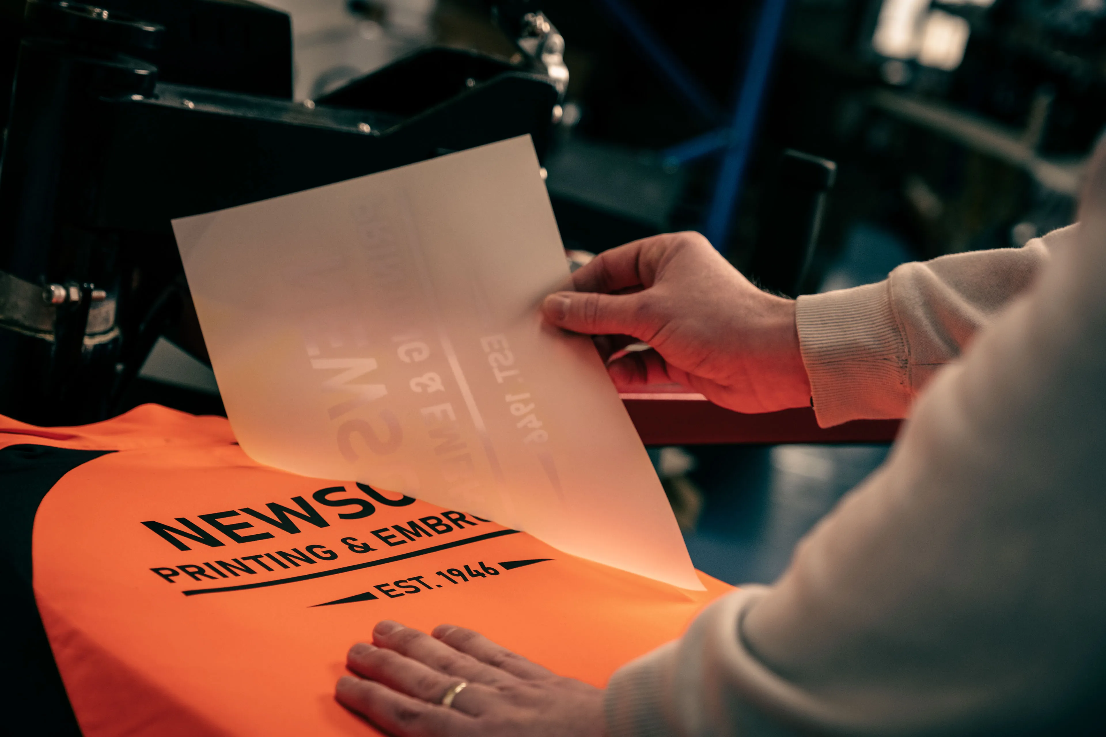 Person in light sweater preparing to apply a transfer onto an orange garment labeled "NEWSONS PRINTING & EMBROIDERY EST. 1946," conveying a focus on craftsmanship.