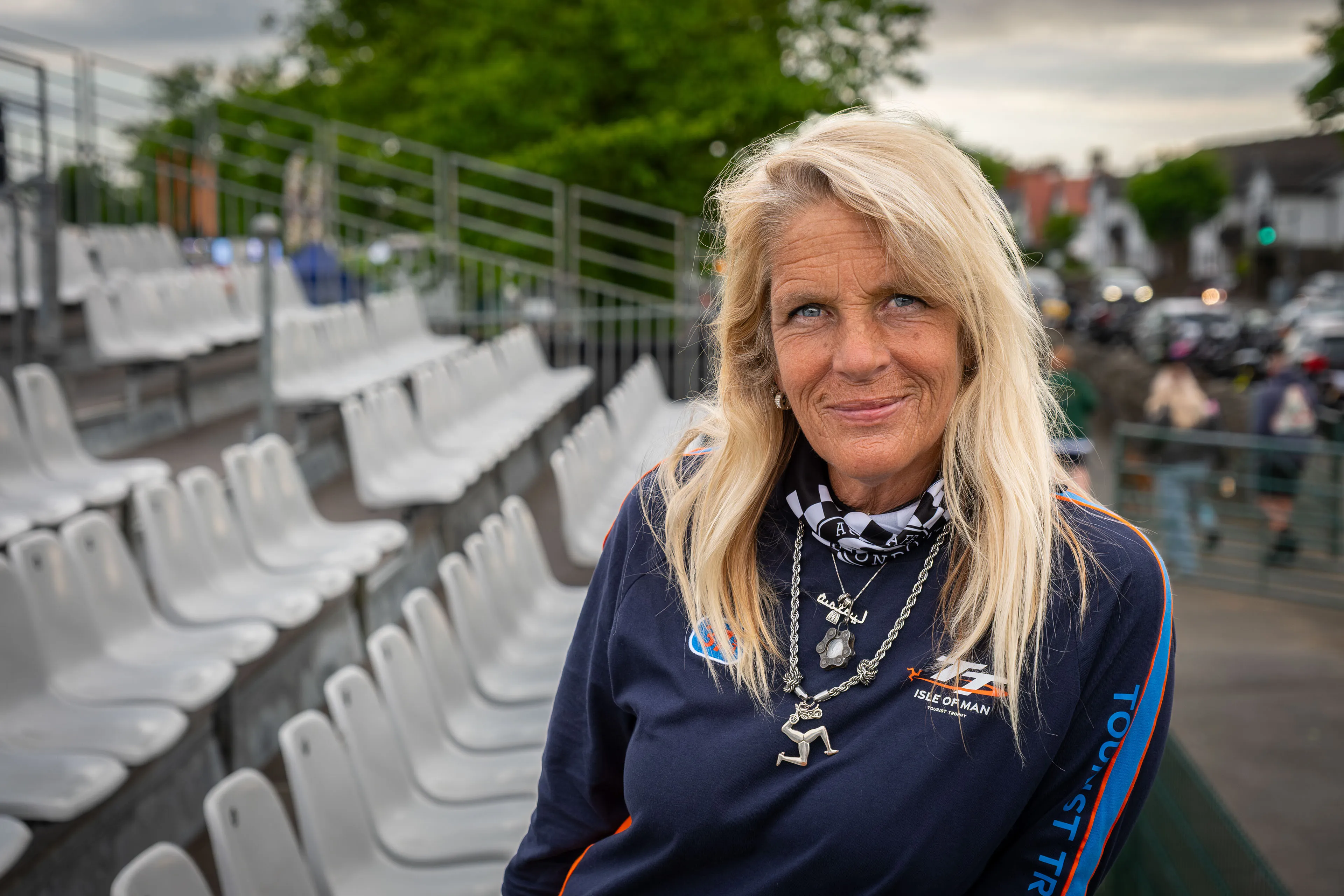 Maria Soledad Facal from Buenos Aires, an Isle of Man TT Marshal, smiling outdoors for Behind the Tabard TT Marshals Series, highlighting marshal stories, in a tt races merch top and collection of silver necklaces