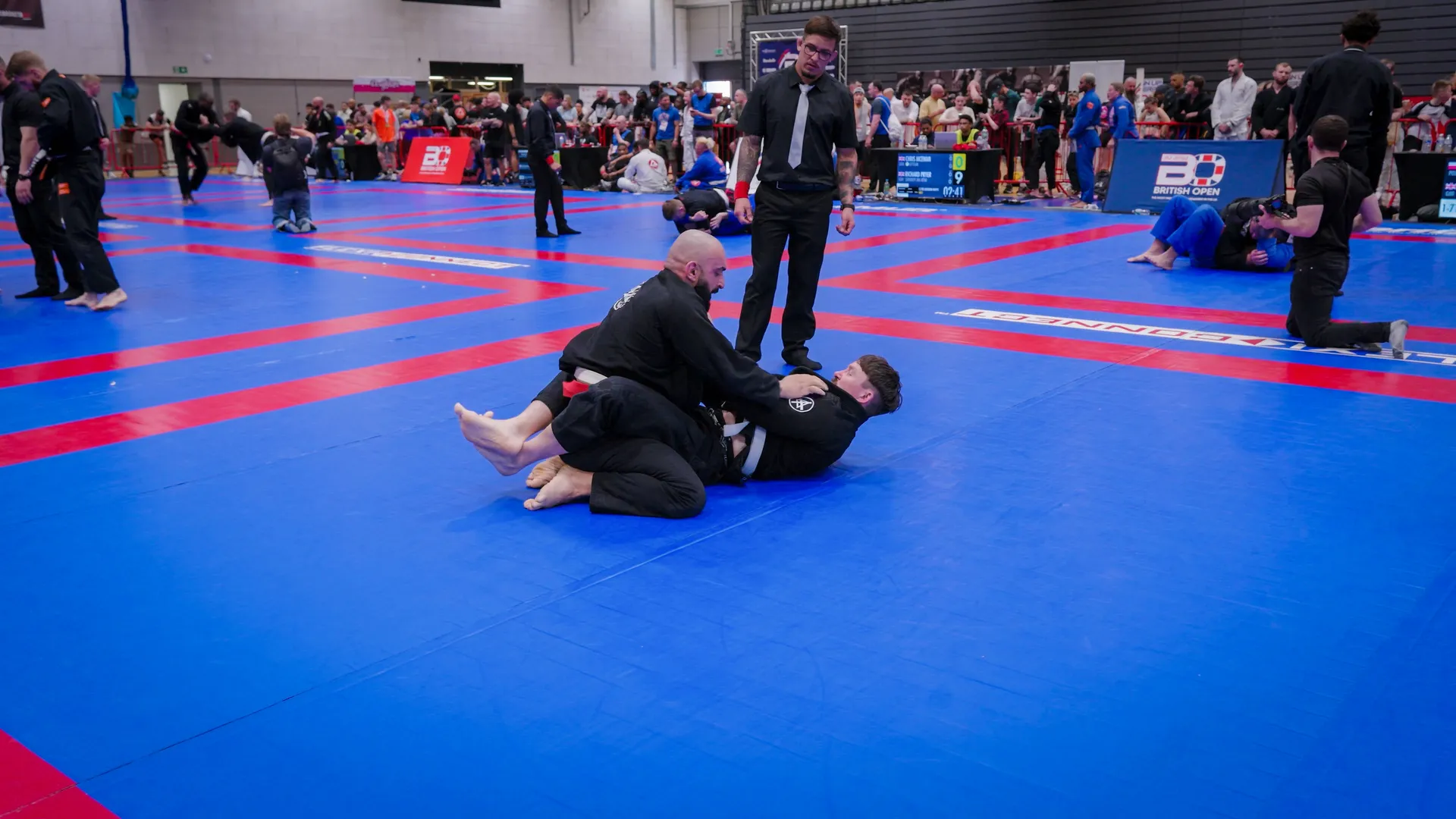 Two competitors in black gis grapple on a blue and red mat in a busy martial arts tournament. A referee observes, with spectators and other matches in the background.