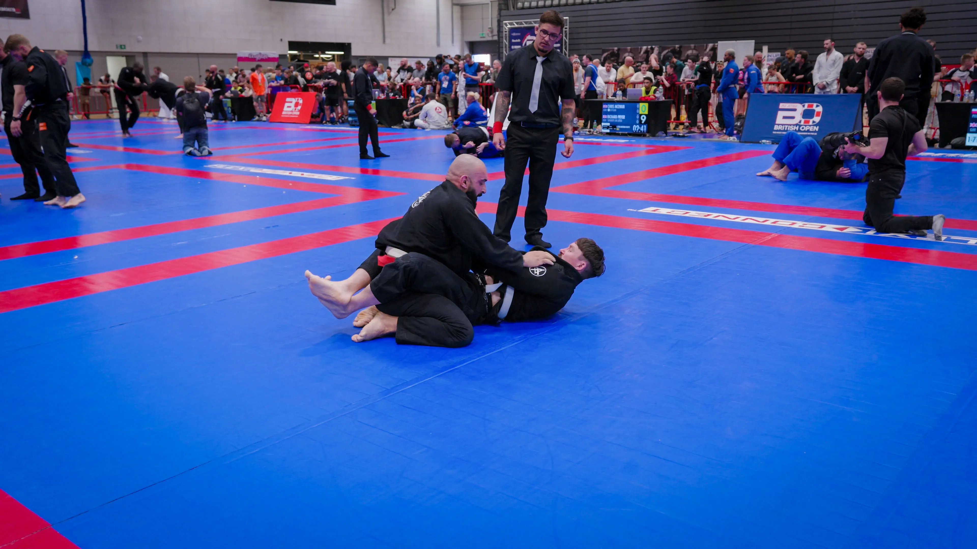Two competitors in black gis grapple on a blue and red mat in a busy martial arts tournament. A referee observes, with spectators and other matches in the background.