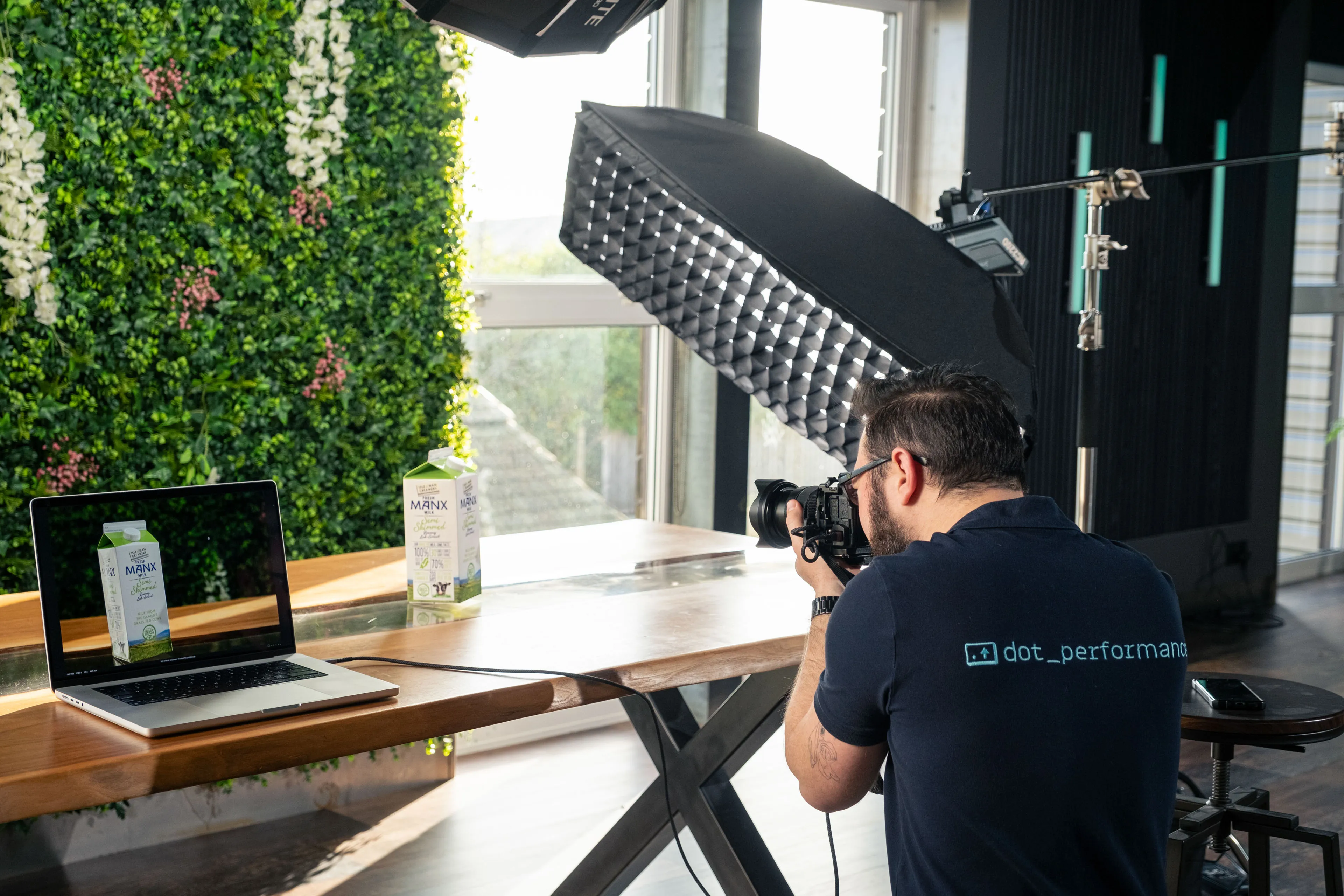 A photographer takes a product shot of a Manx milk carton next to a laptop displaying the product, set against a green living wall and professional softbox lighting.
