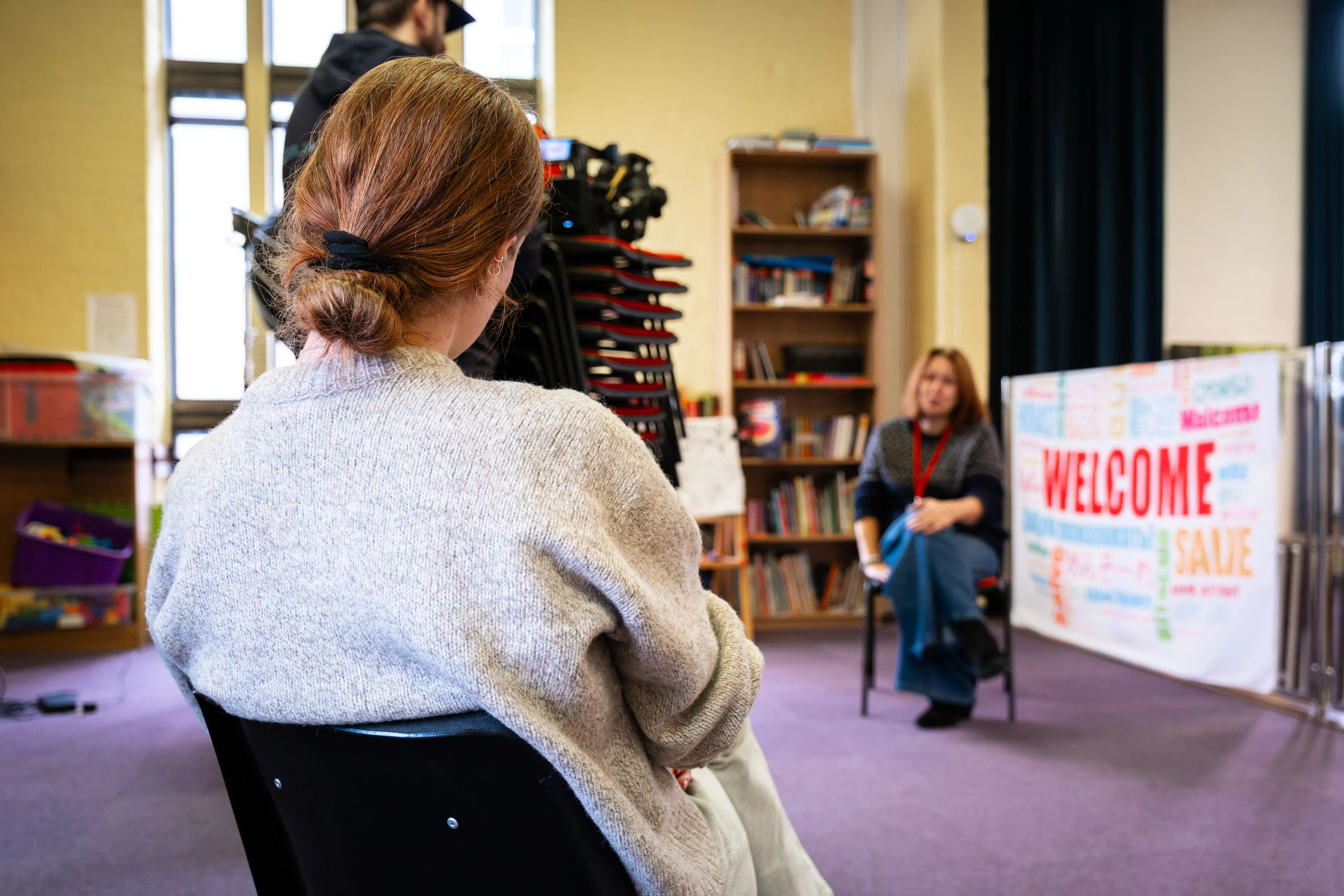 Ella Garvey seated facing the interview setup at Douglas Promenade Church, with Sylvie Géal-Wilkes and a Welcome Project banner visible in the background during documentary filming on the Isle of Man