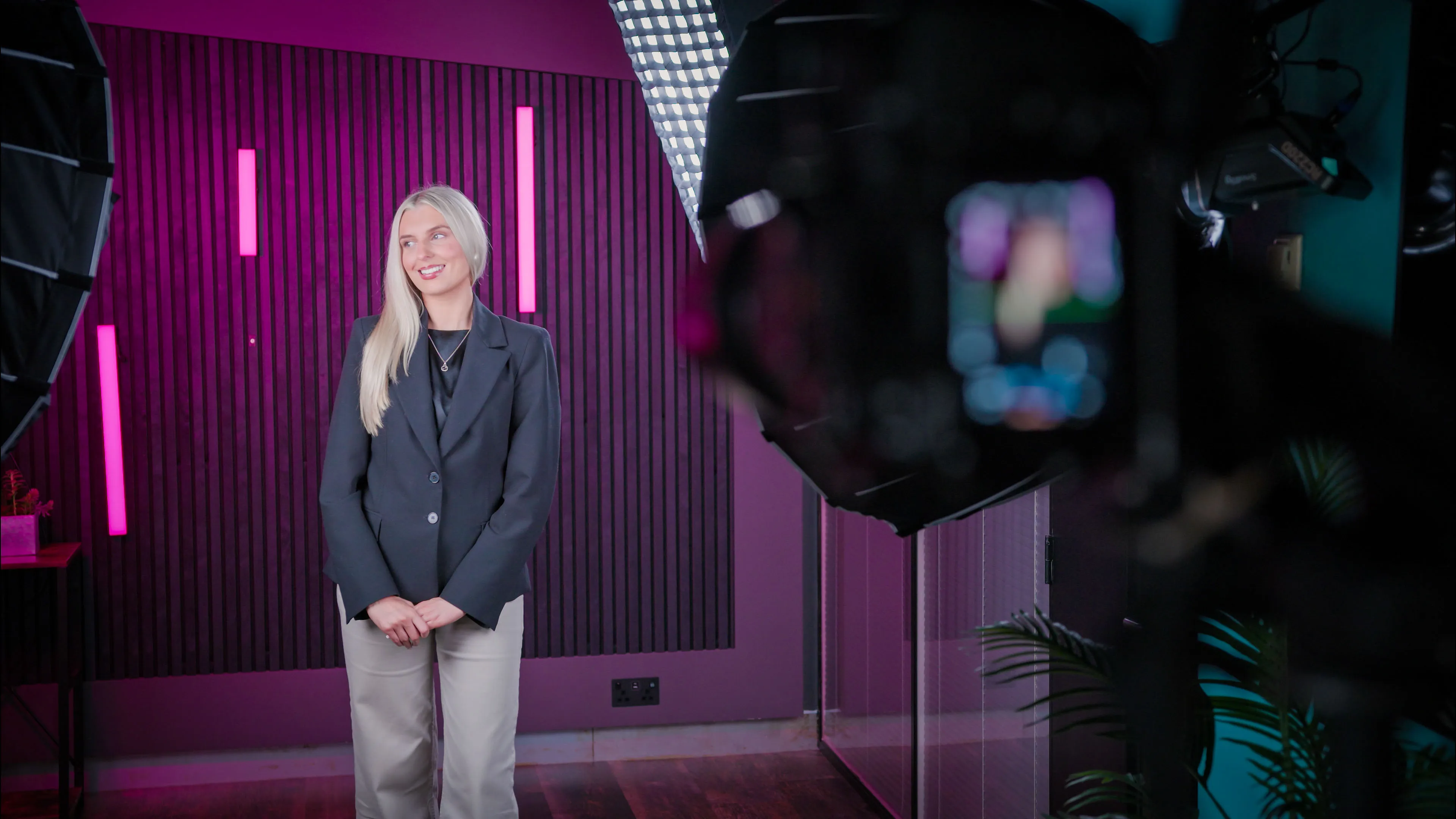 A woman with long blonde hair smiles during a video shoot. She stands in a studio with pink lighting and vertical wall panels, surrounded by camera equipment.