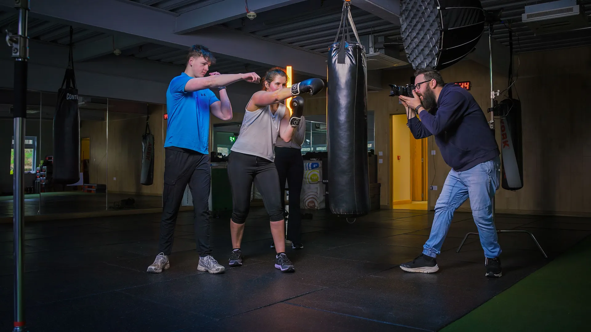 A boxing trainer instructs a woman in gloves, while a photographer captures the scene in a gym. The setting is dynamic and focused, conveying determination.