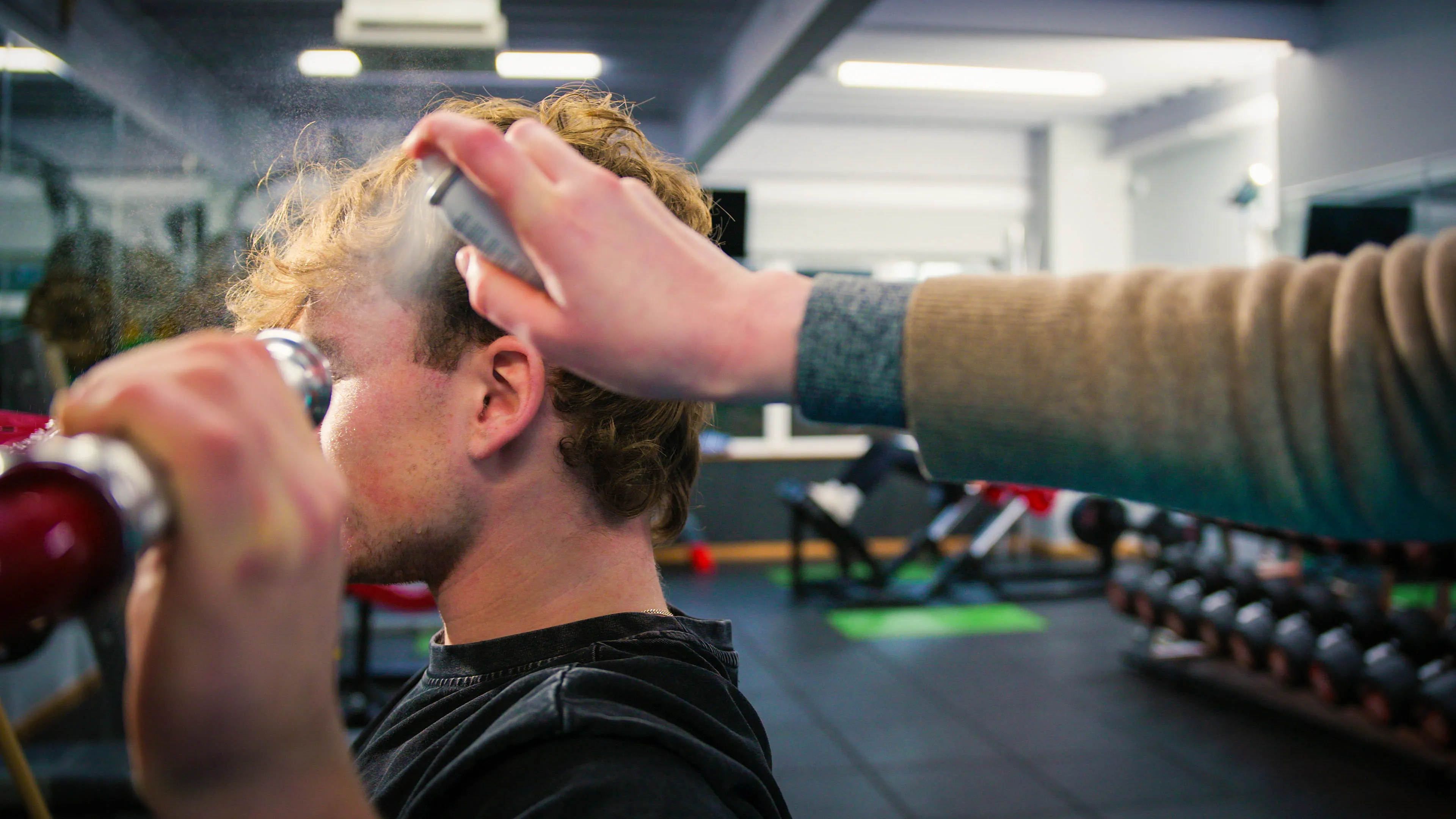 A person with curly hair receives a mist of water spray to emulate sweat, in a bright gym. A hand applies the spray, with exercise equipment blurred in the background.