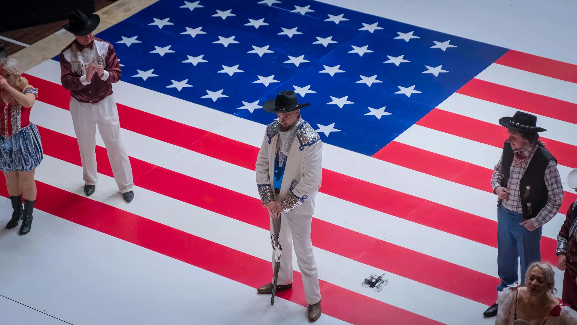 Performers in cowboy attire stand on a large American flag backdrop, conveying a patriotic and Western-themed tone.