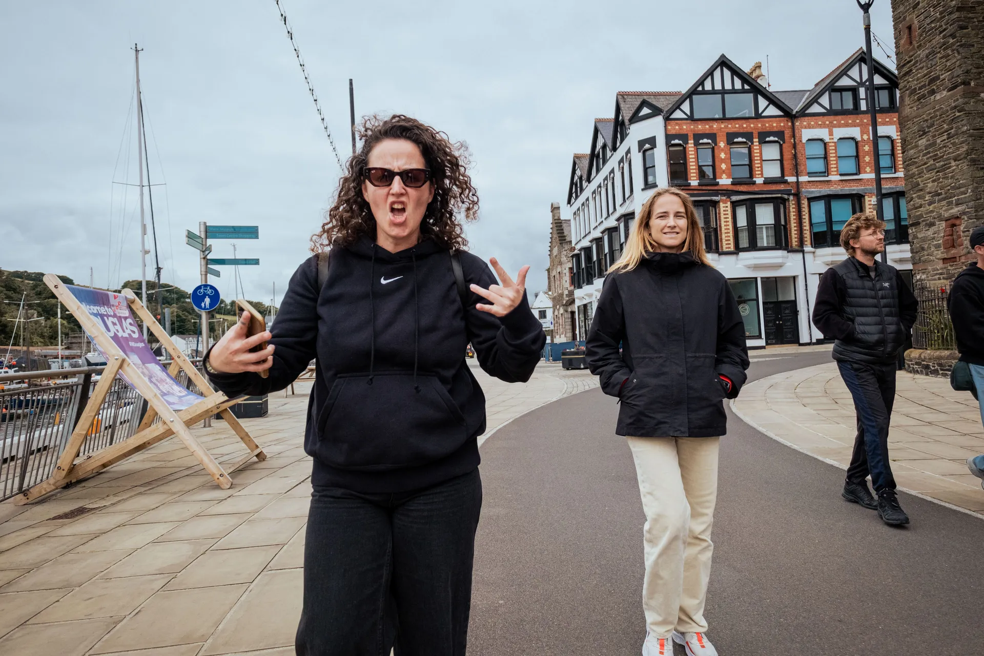 A woman in a black hoodie and sunglasses makes a playful face while holding a phone, walking beside another woman in a black jacket. Two other people stroll in the background, with a historic building and a dock lined with boats under an overcast sky. The mood is lively and casual.