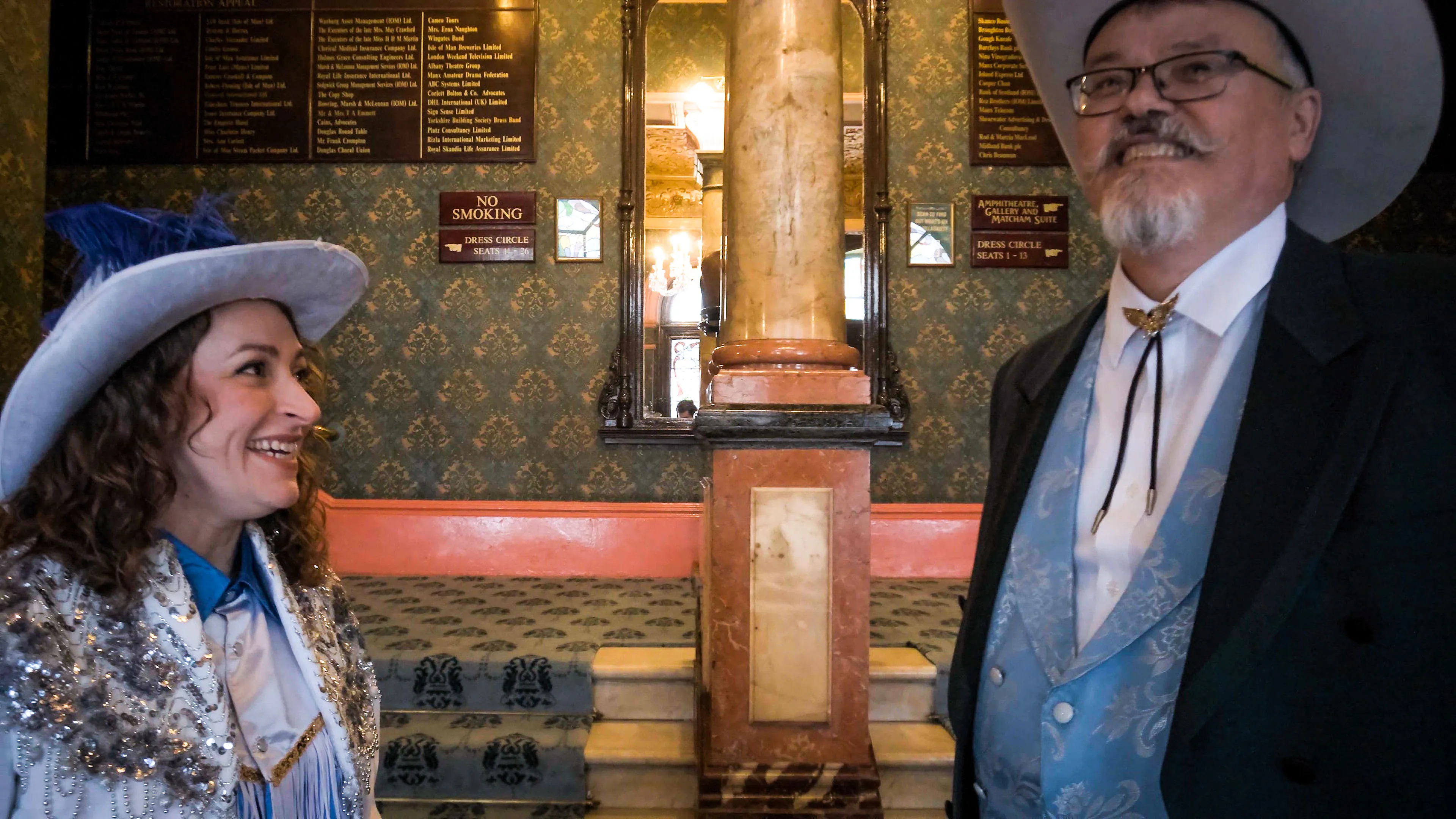 A smiling woman and man in cowboy hats and Western attire stand in a patterned, ornate hallway, evoking a lively and nostalgic Western ambiance.
