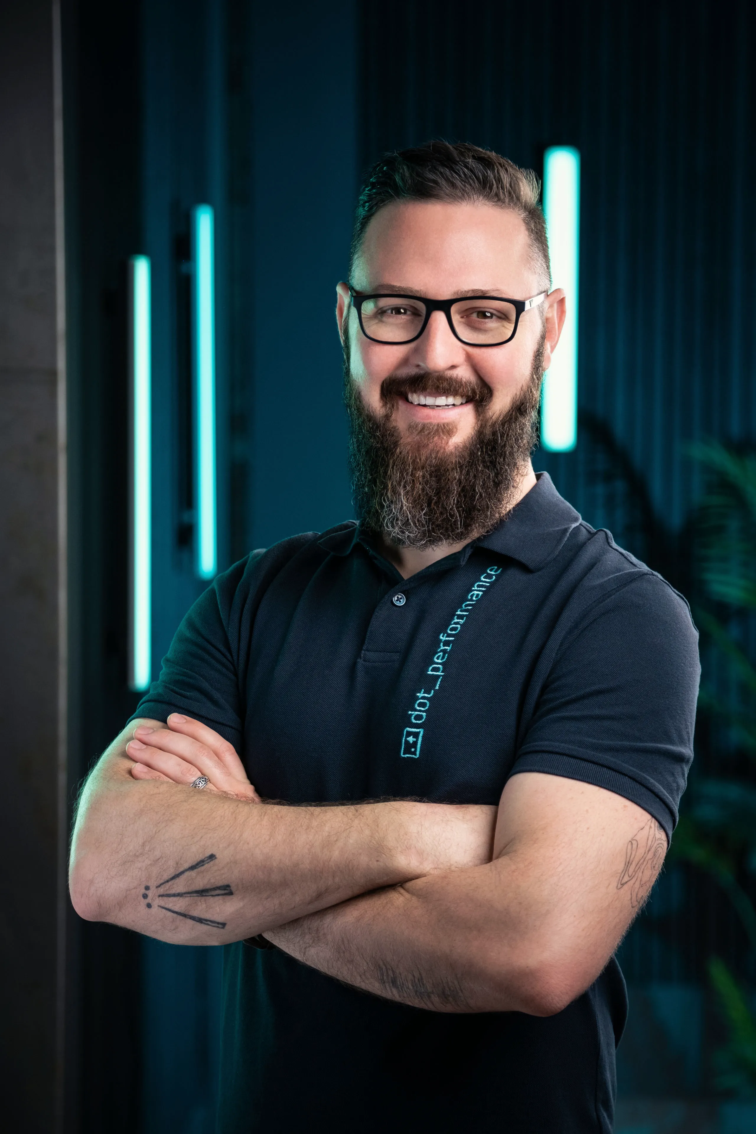 Nico Van Loggerenberg, Photographer and Cinematographer at DotPerformance, smiling with a Beard wearing a DotPerformance branded polo shirt at DotPerformance studios on the Isle of Man, against a dark background with teal neon lighting