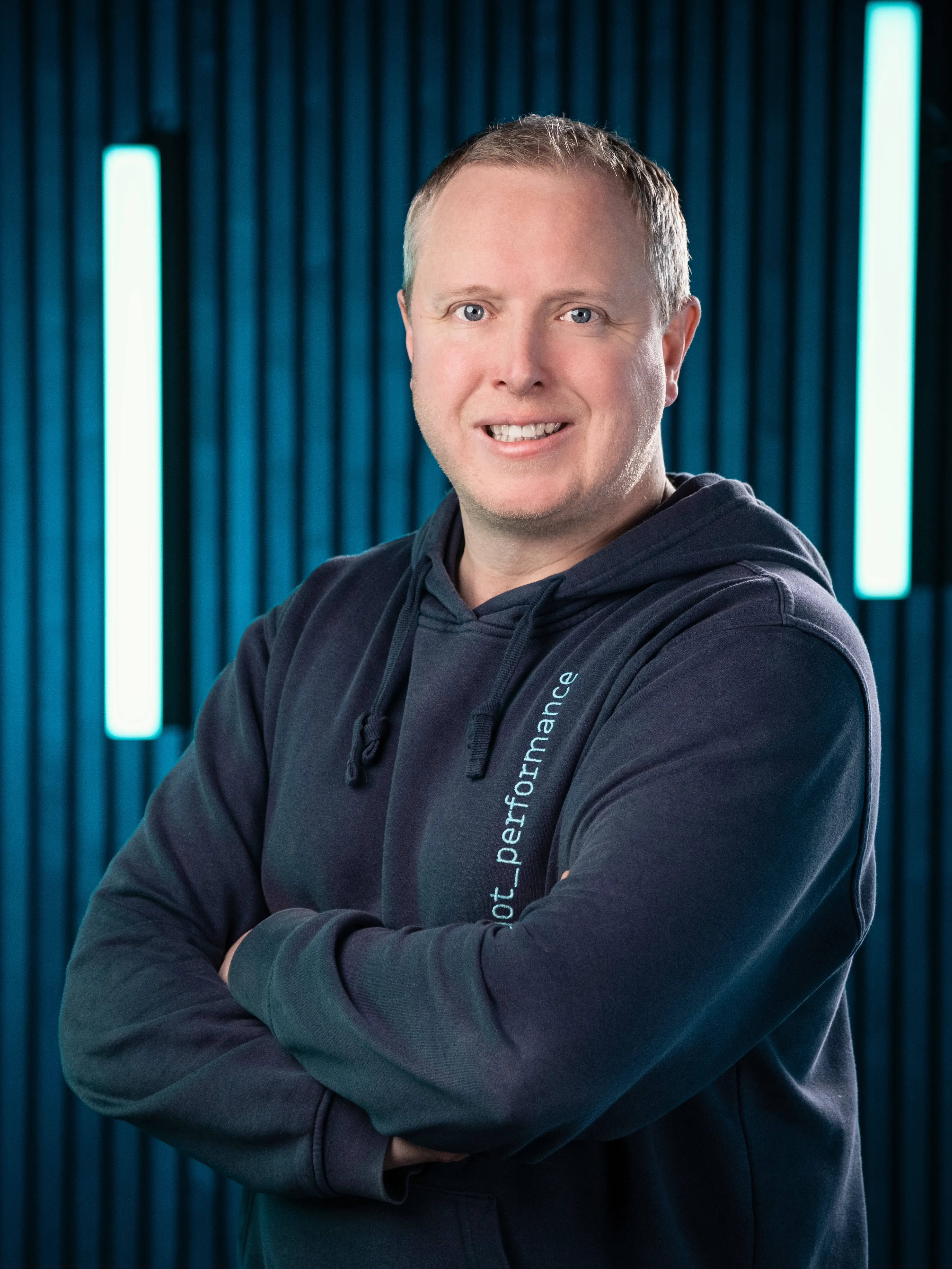 Juan, systems specialist at Dot Performance, smiling with crossed arms as he poses for headshot wearing branded hoody, against dark background with blue neon lights.