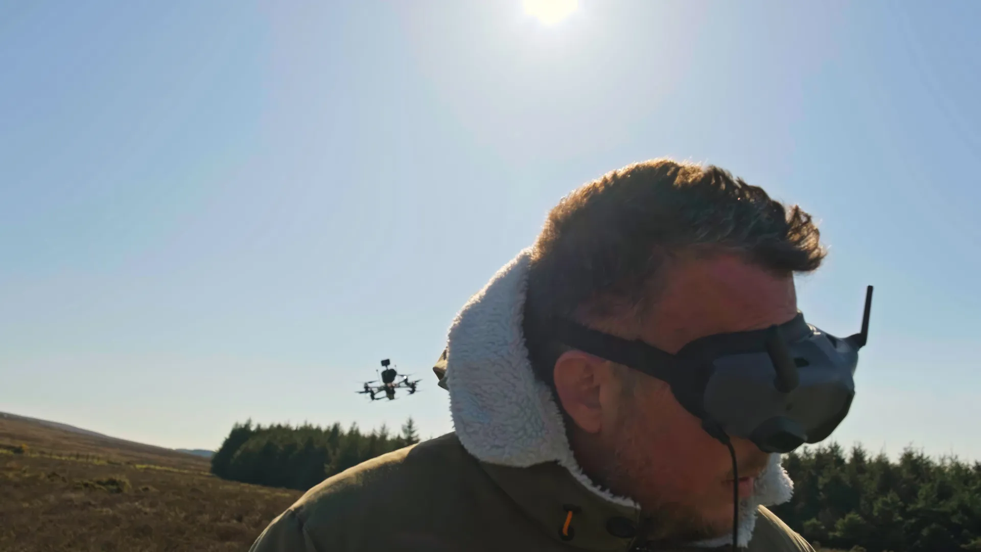 Andy Orton wearing FPV pilot goggles on the Isle of Man with a cinelifter drone visible in flight behind him during a DotPerformance video production shoot