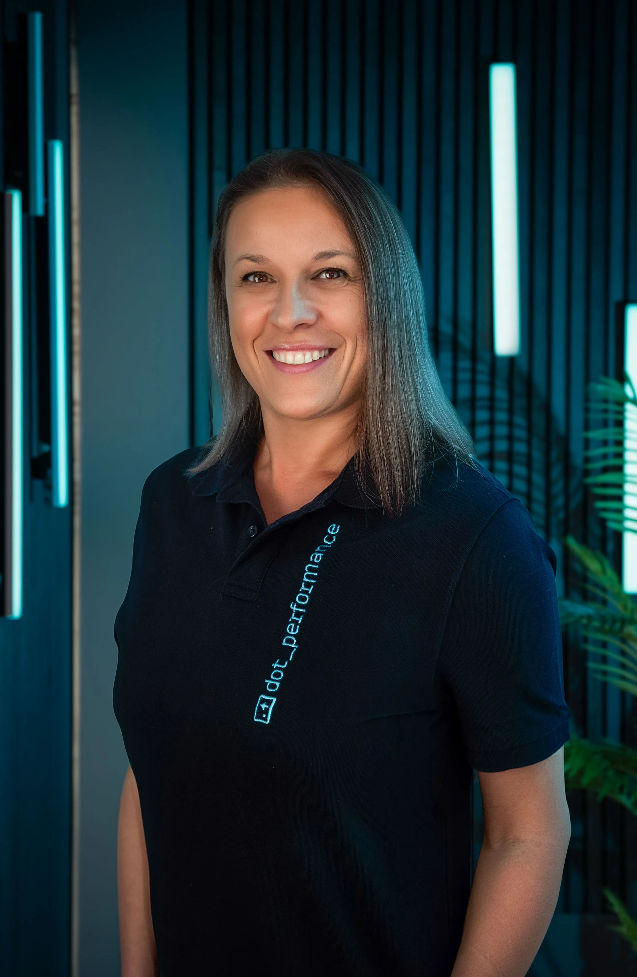Veronica Danila, Front-End Developer at DotPerformance, smiling wearing a DotPerformance branded polo shirt at DotPerformance studios on the Isle of Man, against a dark background with teal neon lighting