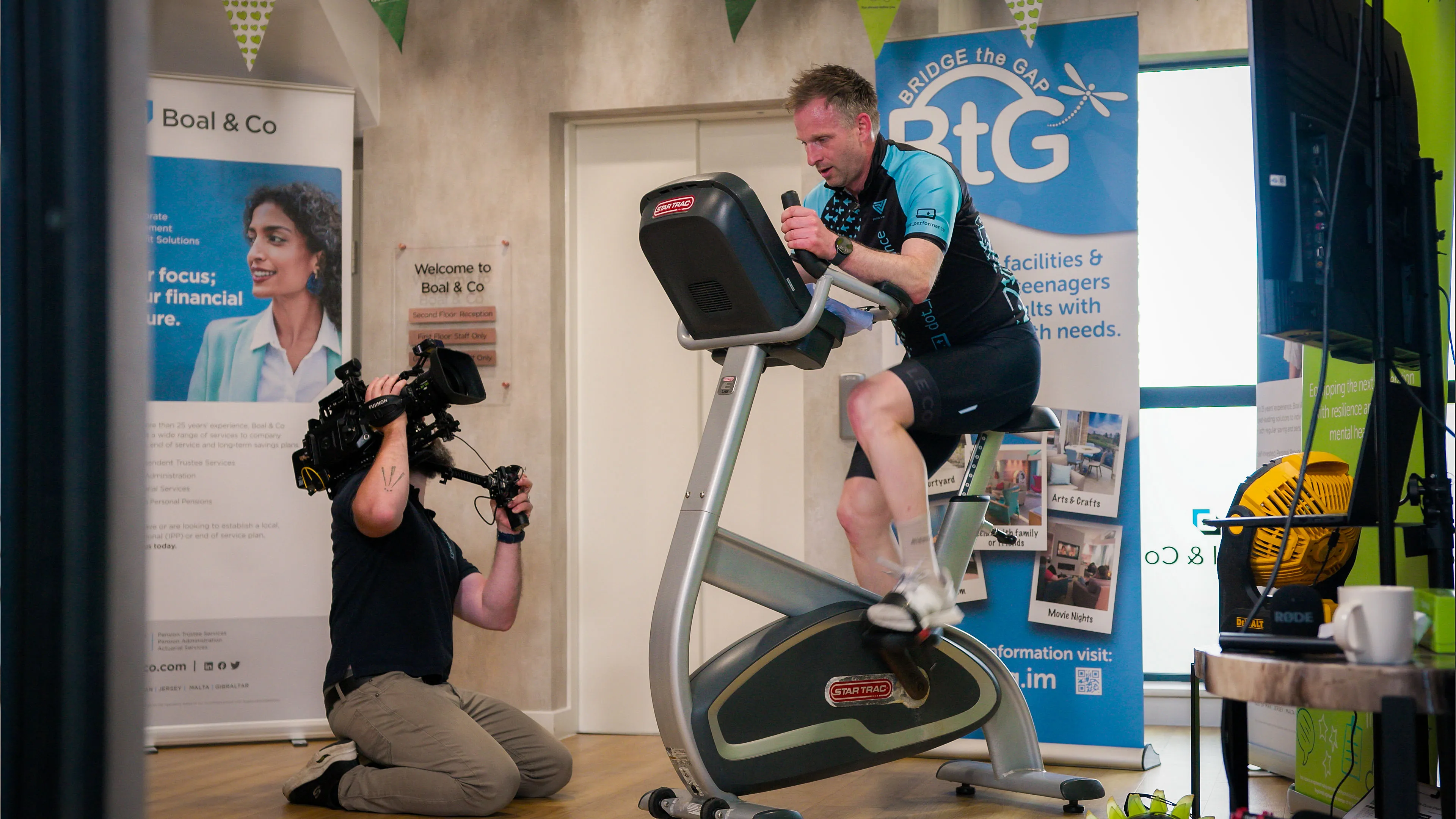 Nico van Loggerenberg from DotPerformance kneeling on the floor to film John Beaty cycling on a static bike with a Blackmagic URSA 12K Mini Pro at the Boal & Co office, Isle of Man.