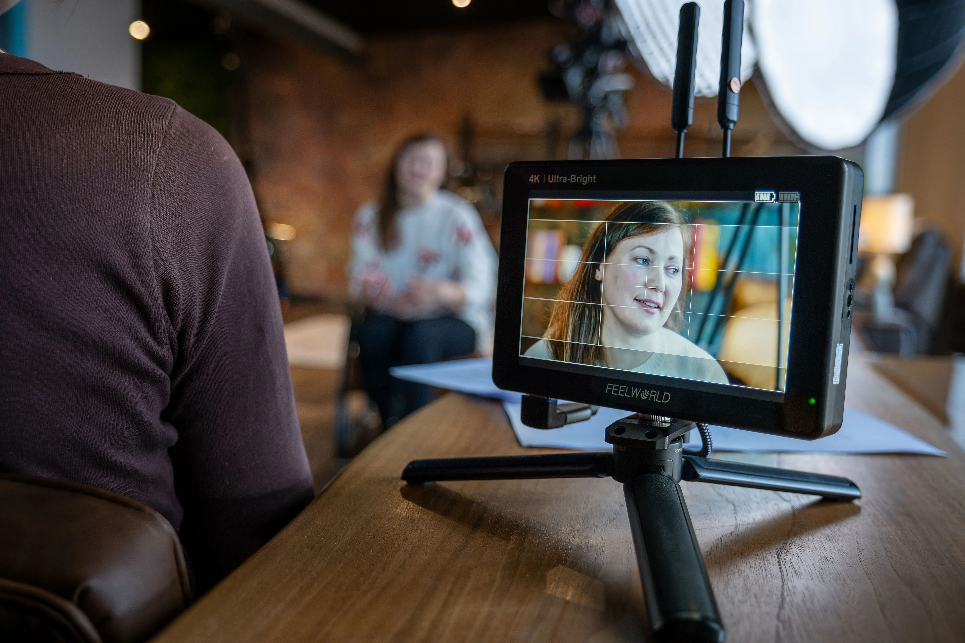 A woman being interviewed on camera, visible on a monitor screen in focus. The setting is cozy, with warm lighting and a blurred background.