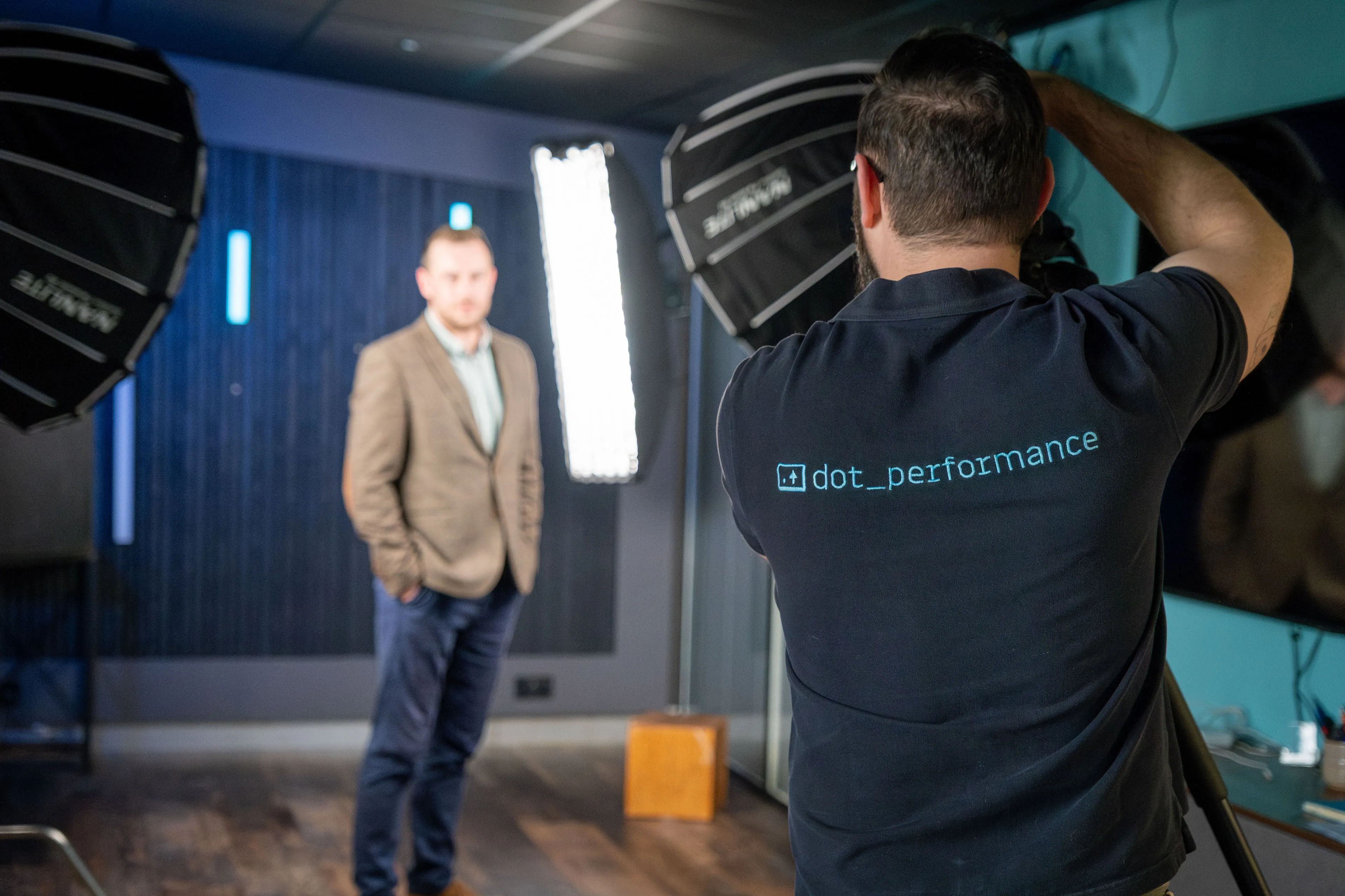 A photographer in a blue shirt photographs a man in a blazer under studio lighting. The setting is professional, with a calm, focused ambiance.