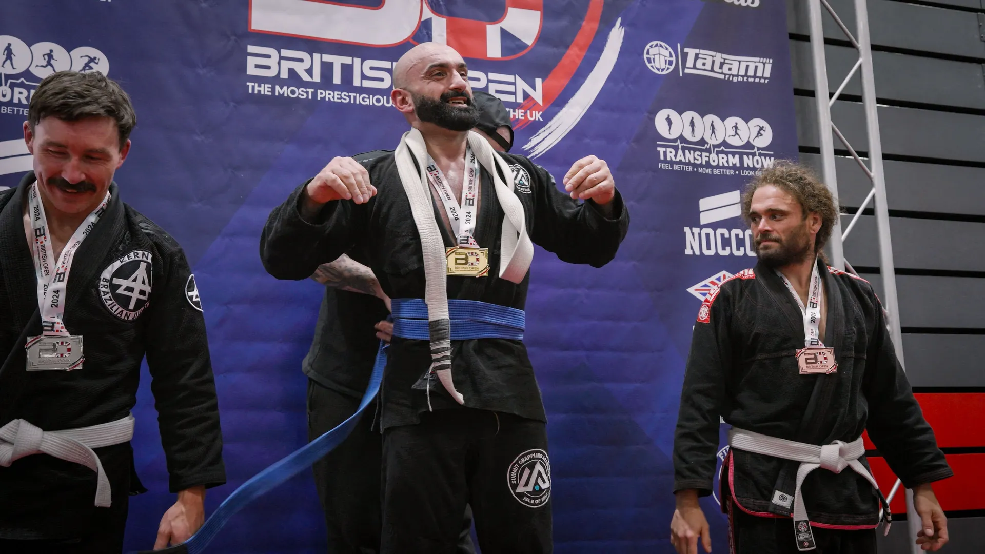 Three men stand on a podium at a Brazilian Jiu-Jitsu competition, all in black gis, wearing medals. The center man smiles broadly, exuding joy.