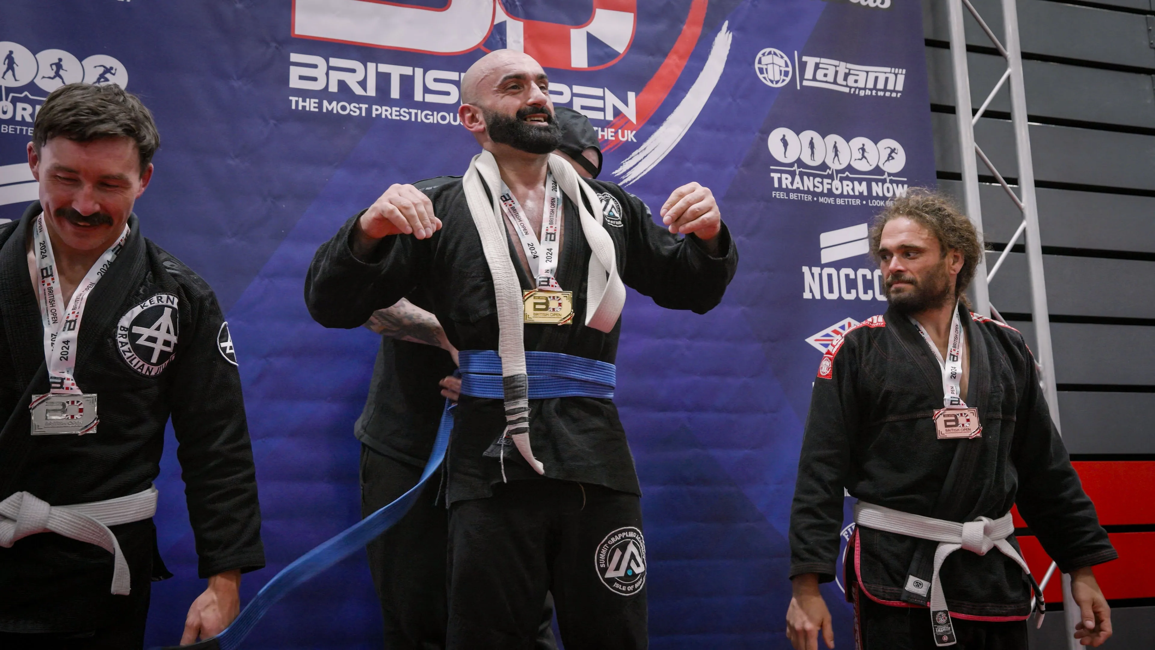 Three men stand on a podium at a Brazilian Jiu-Jitsu competition, all in black gis, wearing medals. The center man smiles broadly, exuding joy.