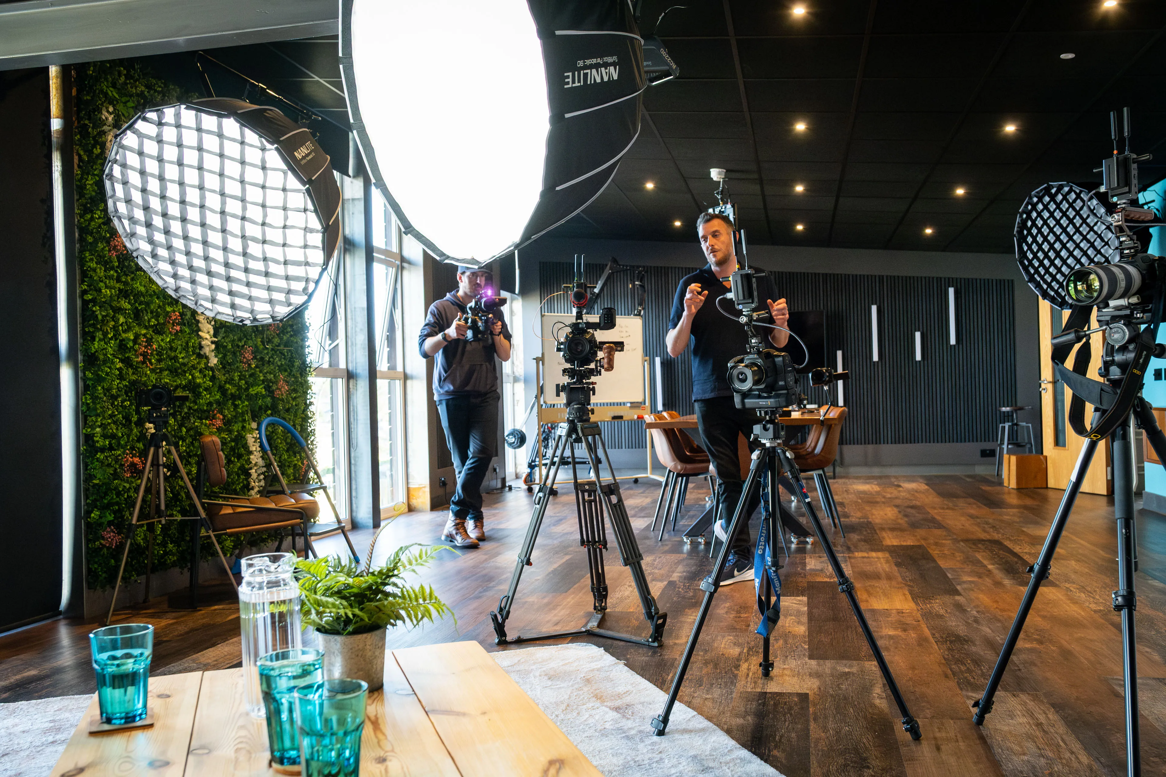 Andy Orton and Will Oates operating cinema cameras and adjusting lighting rigs in a studio with a living wall and large windows during a multi-camera podcast production on the Isle of Man