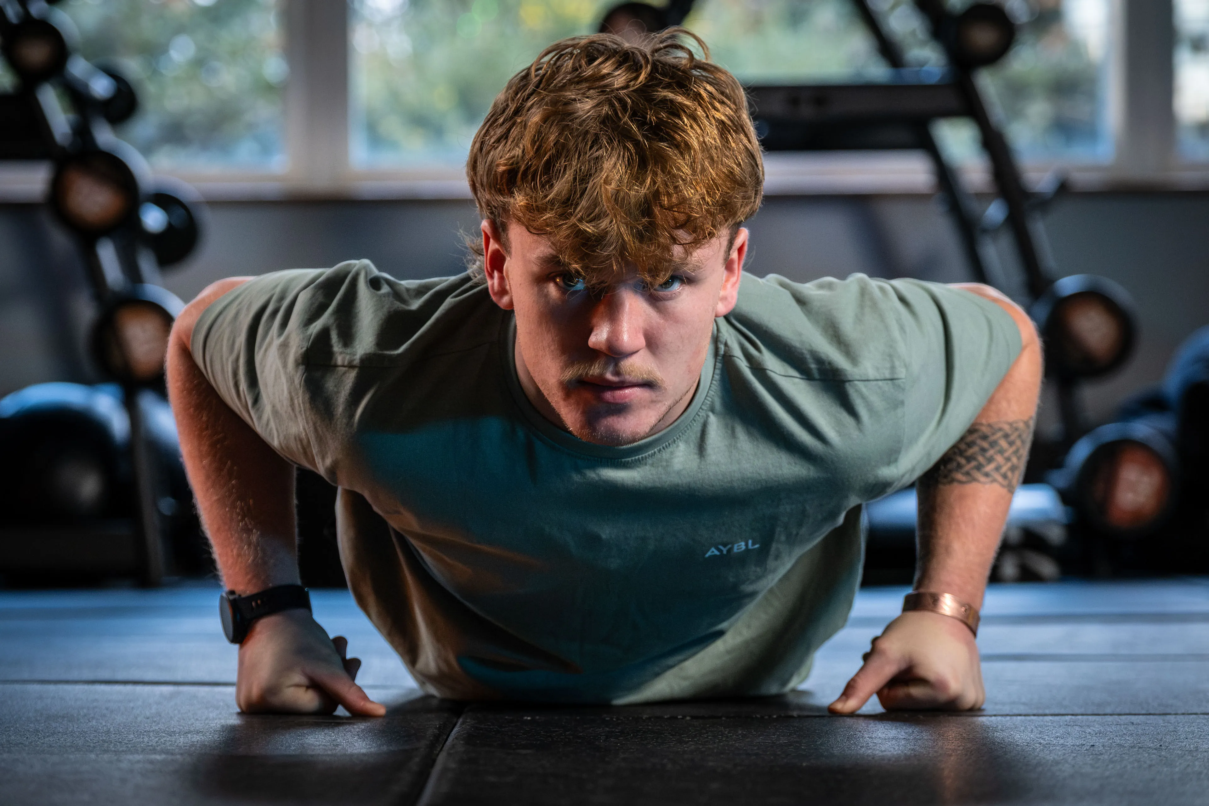Focused young man doing push-ups in a gym, wearing a green shirt. Background features weights and equipment, creating an intense workout atmosphere.