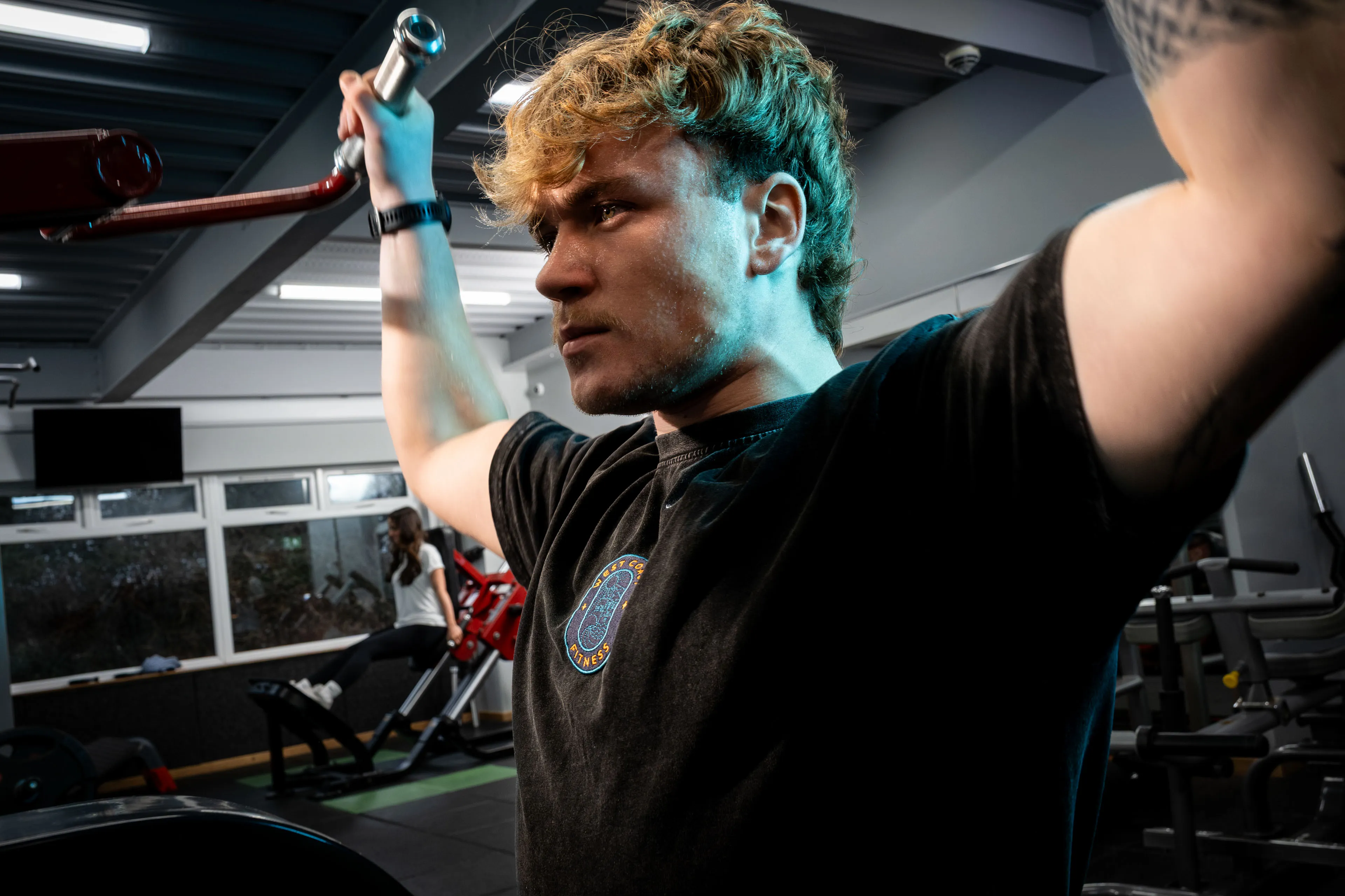 A person with curly hair intensely lifts a barbell in a gym, expressing determination. The room is well-lit, with exercise equipment in the background.