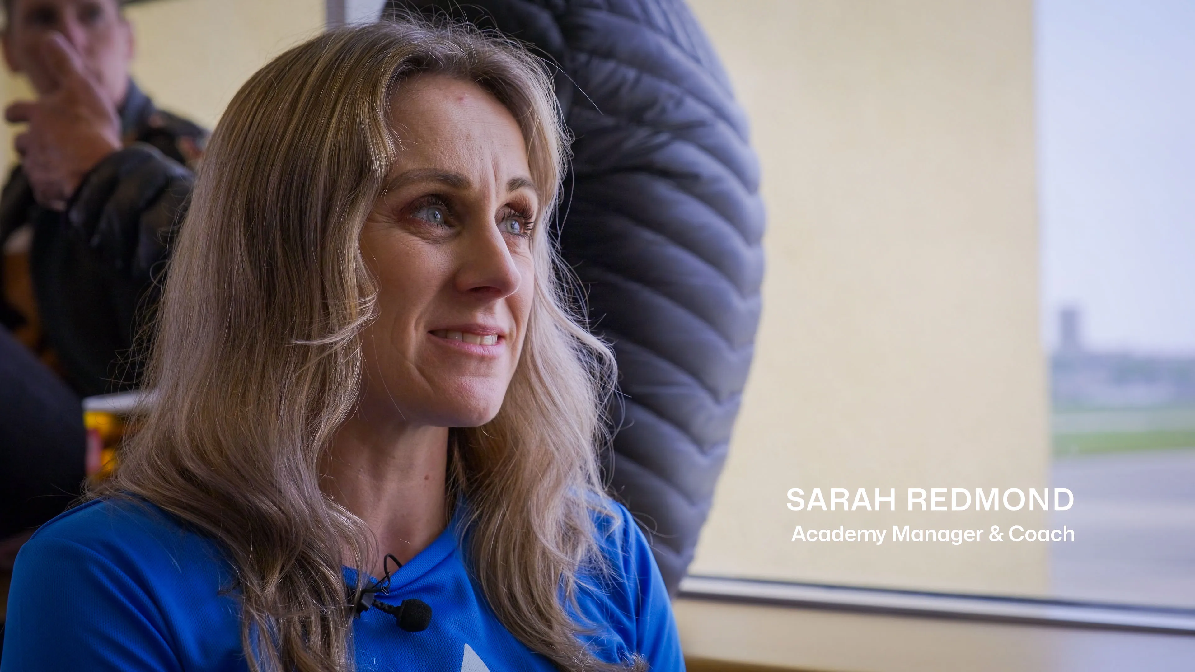 A woman with long hair and a light jacket smiles warmly in a bright room. Text reads "Academy Manager & Coach." A blurred person and window are in the background.