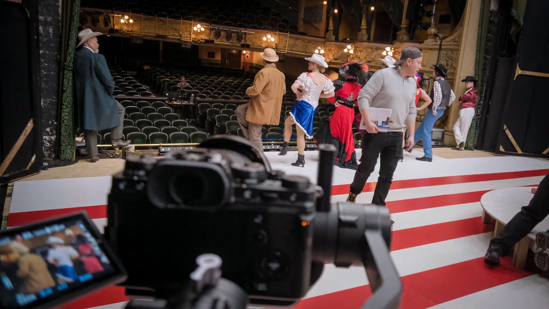 A theater rehearsal with actors in colourful, western-style costumes gathers on a stage. A camera records the scene, capturing lively ambiance.