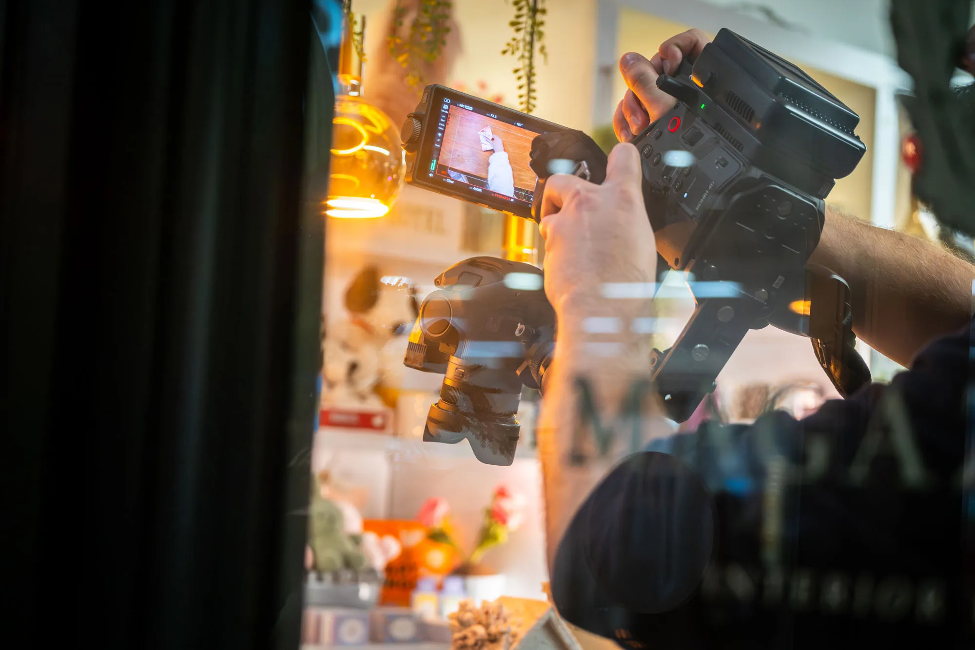A person films through glass with a camera rig, capturing a brightly lit indoor scene with hanging plants and decorations. The atmosphere feels lively and creative.