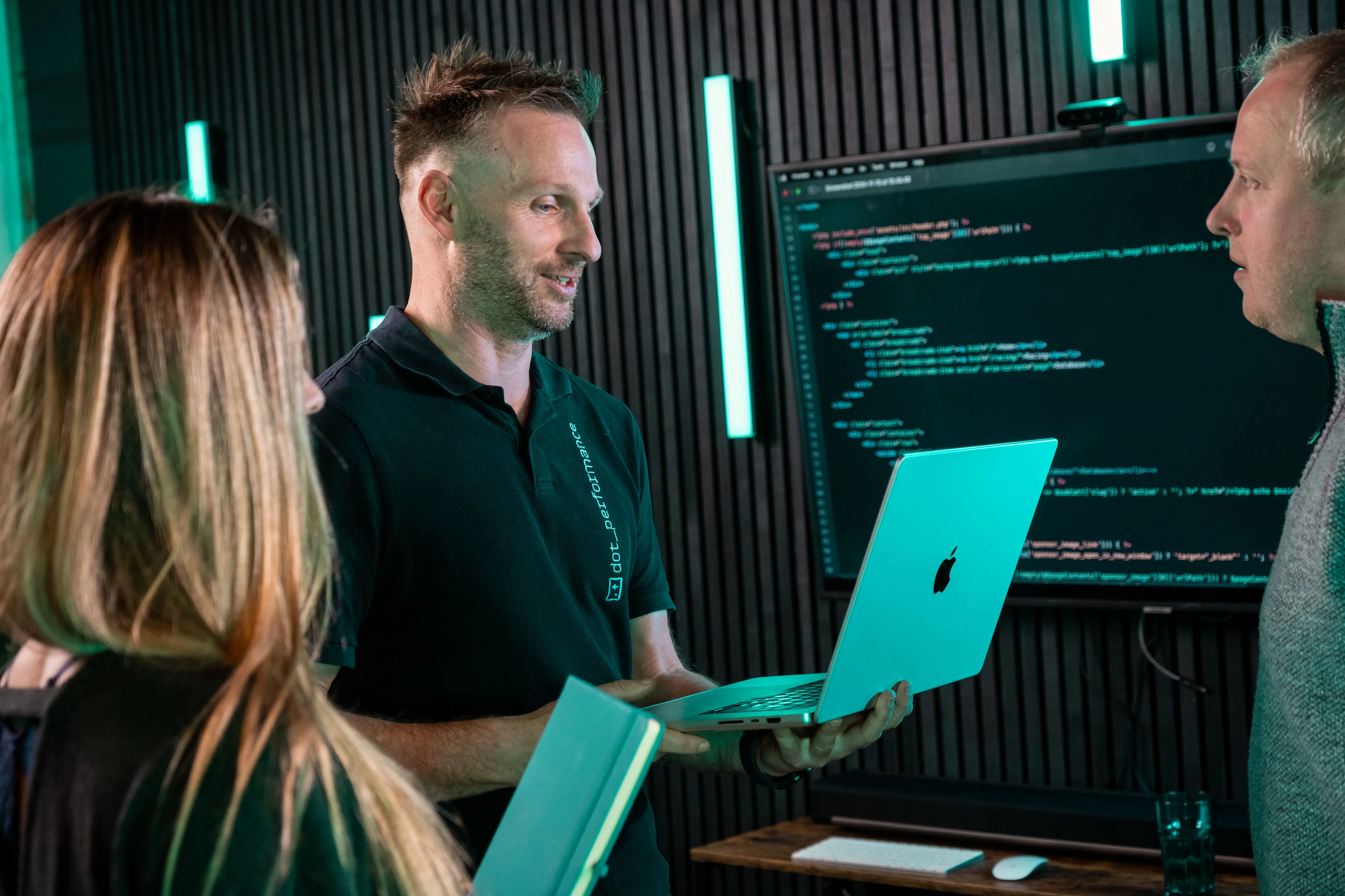 John Beaty holding an Apple MacBook and presenting development work to DotPerformance colleagues Rachael Fabrizio and Juan Readshaw at the agency office in Douglas, Isle of Man, with front-end code displayed on a large screen behind the group