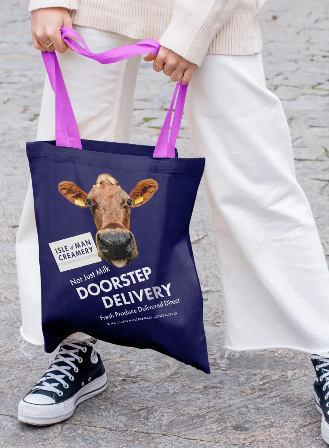 A person wearing white wide-leg trousers and Converse trainers holding a navy tote bag with purple handles, printed with a Jersey cow's face, the Isle of Man Creamery logo, and white text reading "Not Just Milk – Doorstep Delivery – Fresh Produce Delivered Direct"