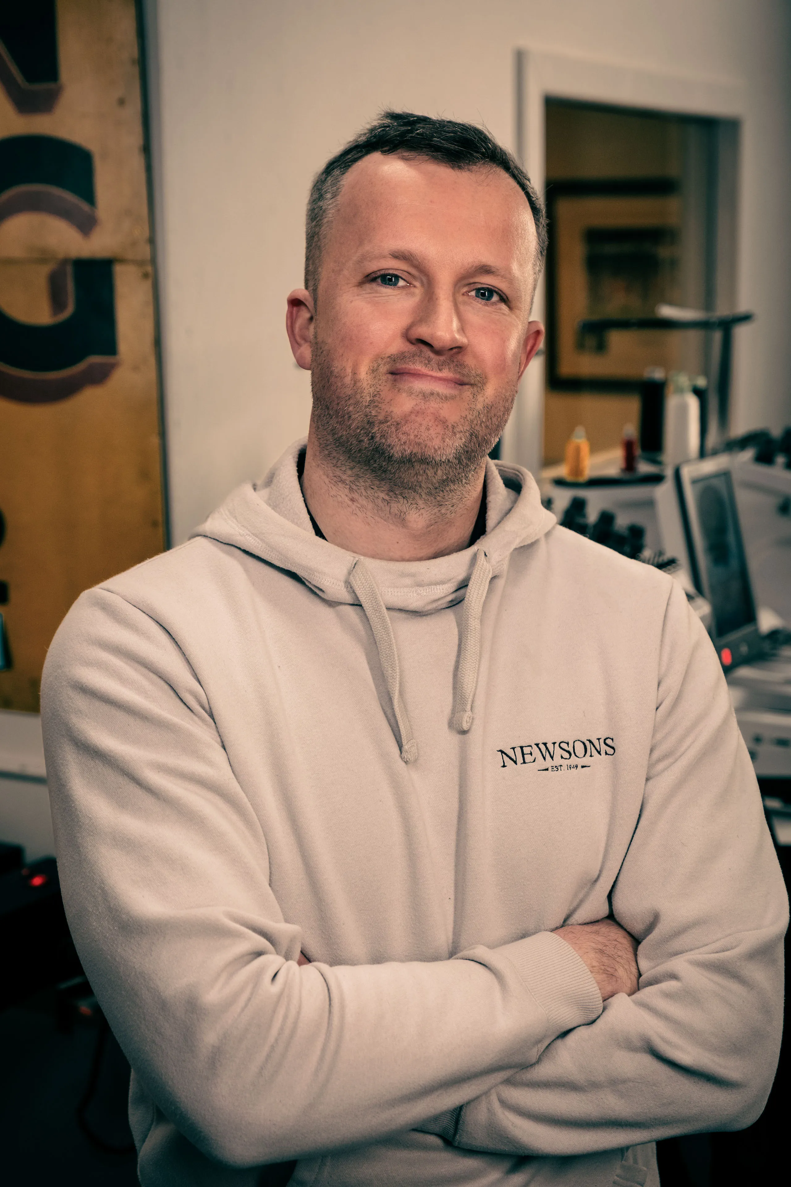 Man in a light hoodie with "NEWSONS" logo, smiling and crossing arms in a cozy room with a warm tone and assorted objects in the background, suggesting a relaxed atmosphere.